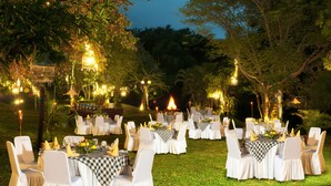 An outdoor dining area with round tables and white chairs, surrounded by trees and string lights. - Prime Plaza Hotel - Purwakarta (Cikampek)