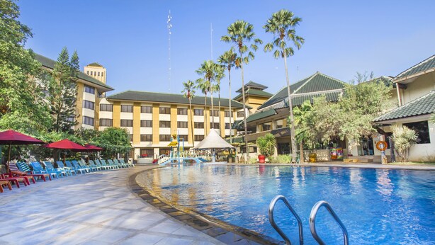 A swimming pool with lounge chairs and umbrellas, surrounded by palm trees and a hotel building. - Prime Plaza Hotel - Purwakarta (Cikampek)