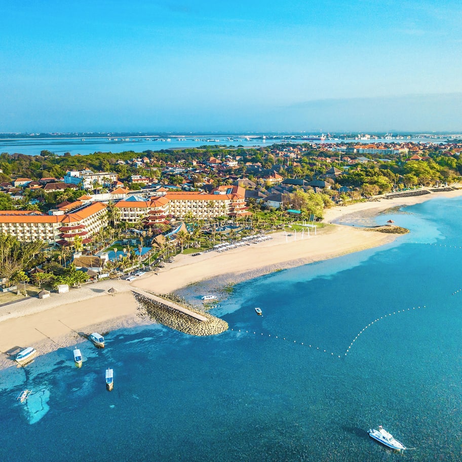 An aerial view of a coastal resort with a beachfront, clear blue waters, and a large hotel complex.