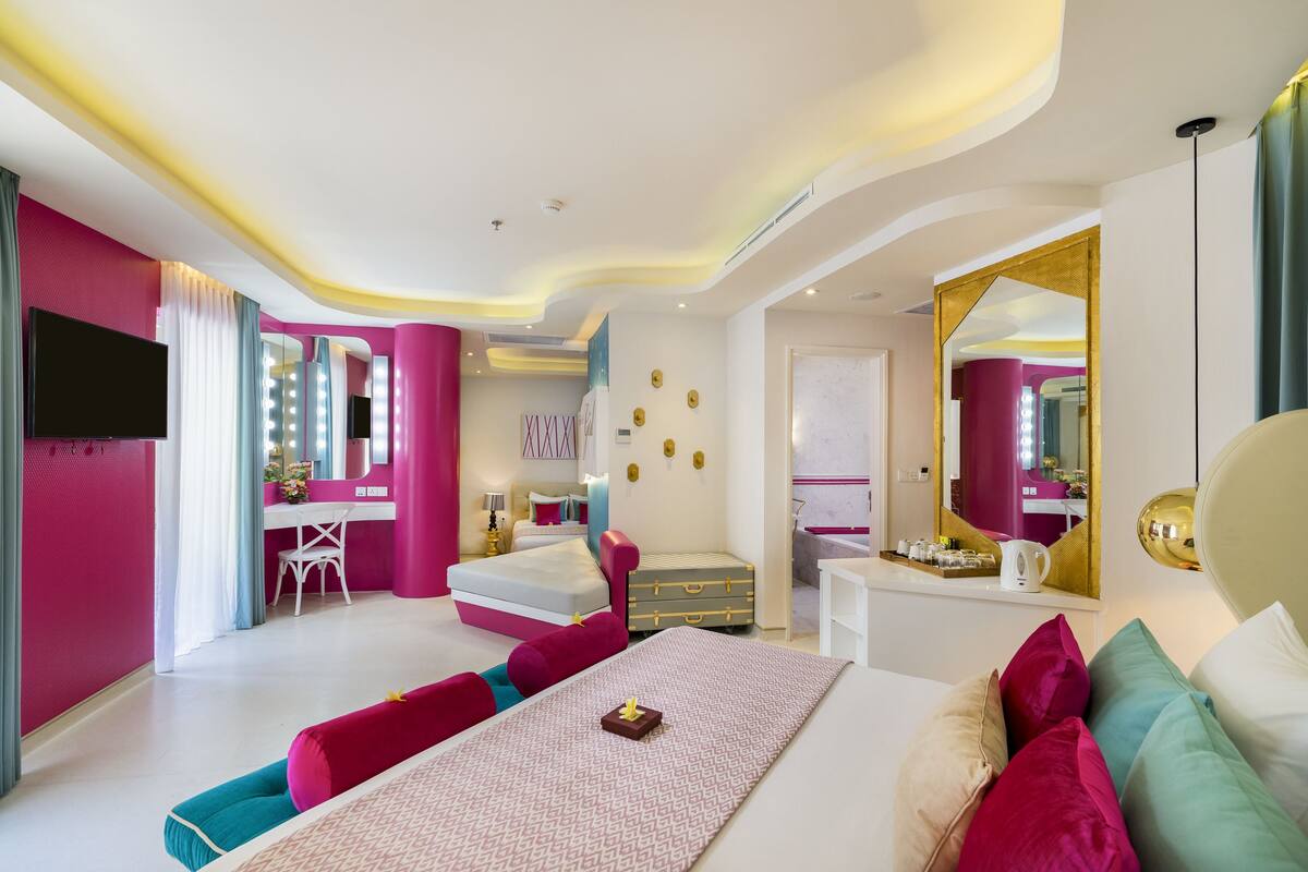 Une chambre d’hôtel moderne aux couleurs rose et blanc, avec un grand lit, une coiffeuse avec éclairage, un bureau et un miroir.
