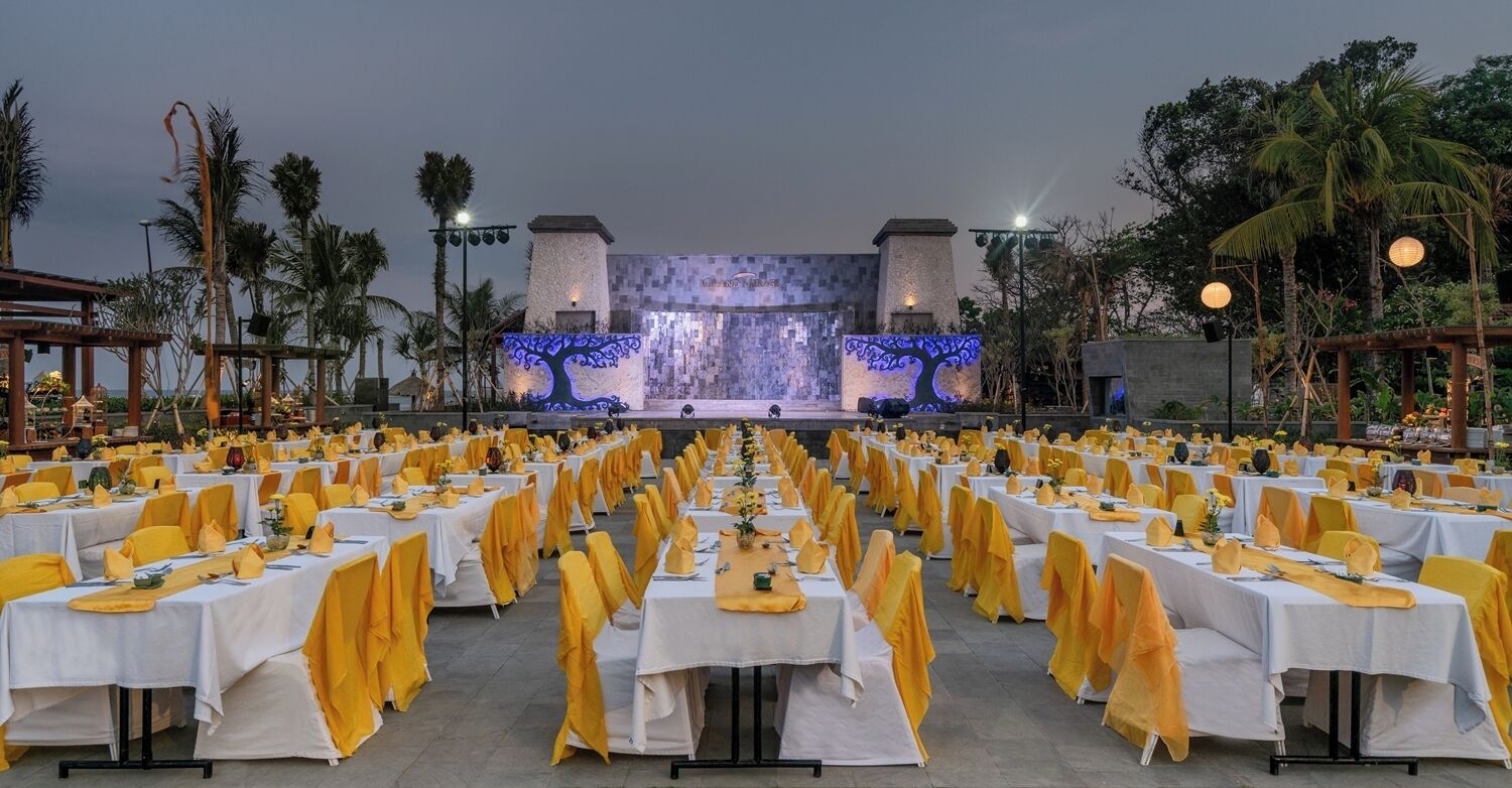 Una sala de banquets a l'aire lliure amb files de taules preparades per a un esdeveniment formal, amb un escenari i una projecció d'un arbre al fons.