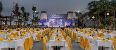 An outdoor banquet hall with rows of tables set for a formal event, featuring a stage with a tree projection in the background.