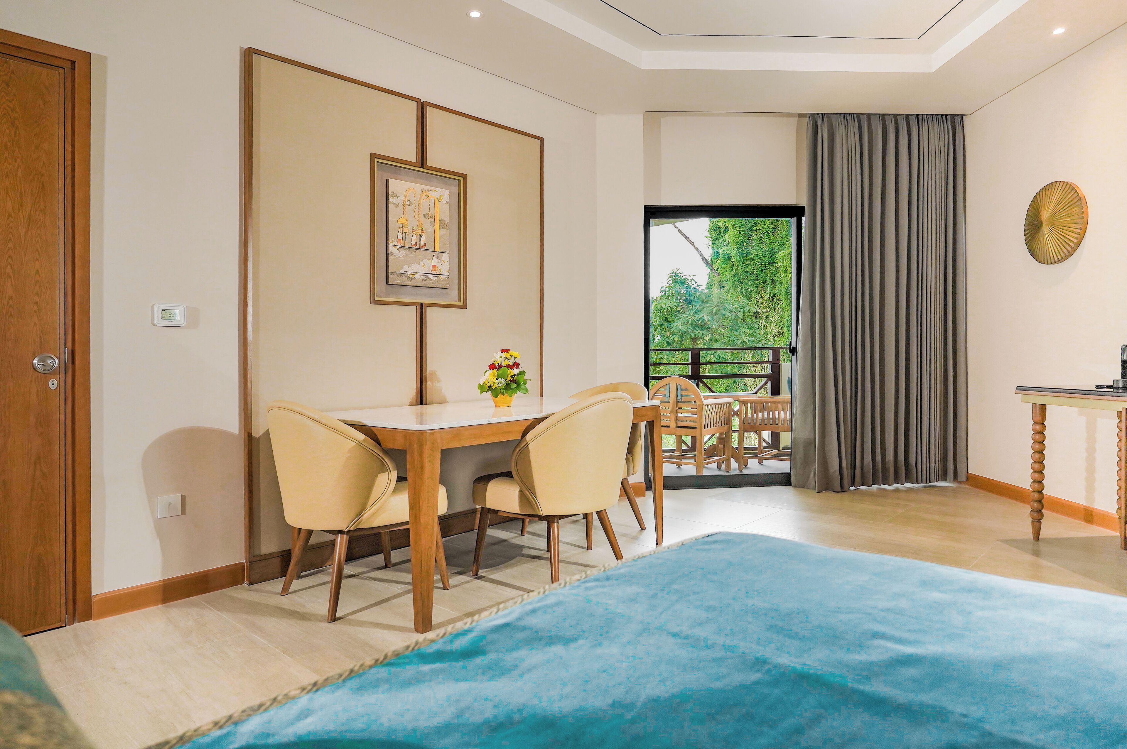 A hotel room with a bed, a dining table with chairs, a small table with a plant, a picture on the wall, and a balcony with a view of greenery.