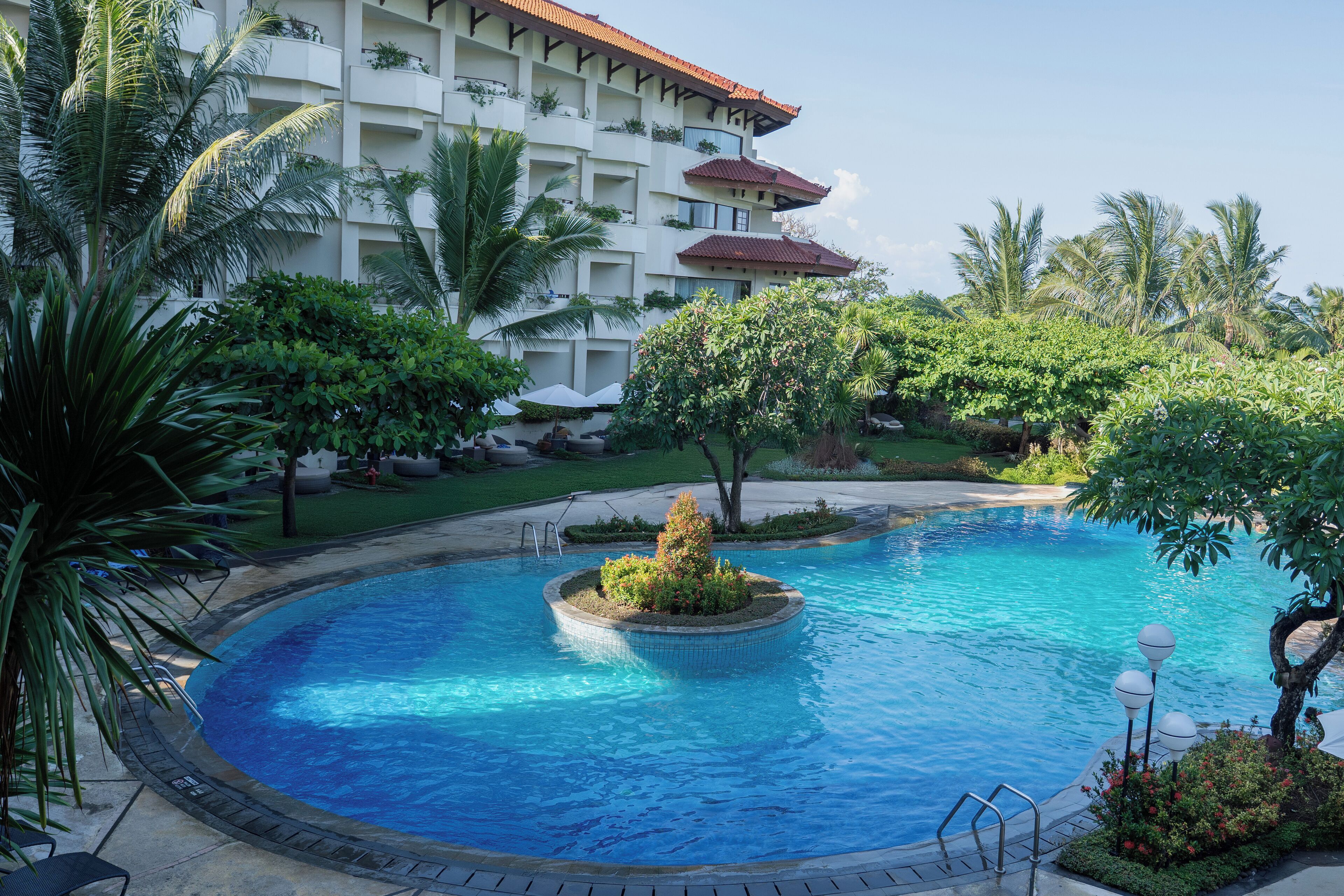 A swimming pool surrounded by lush greenery and a multi-story hotel building with balconies.