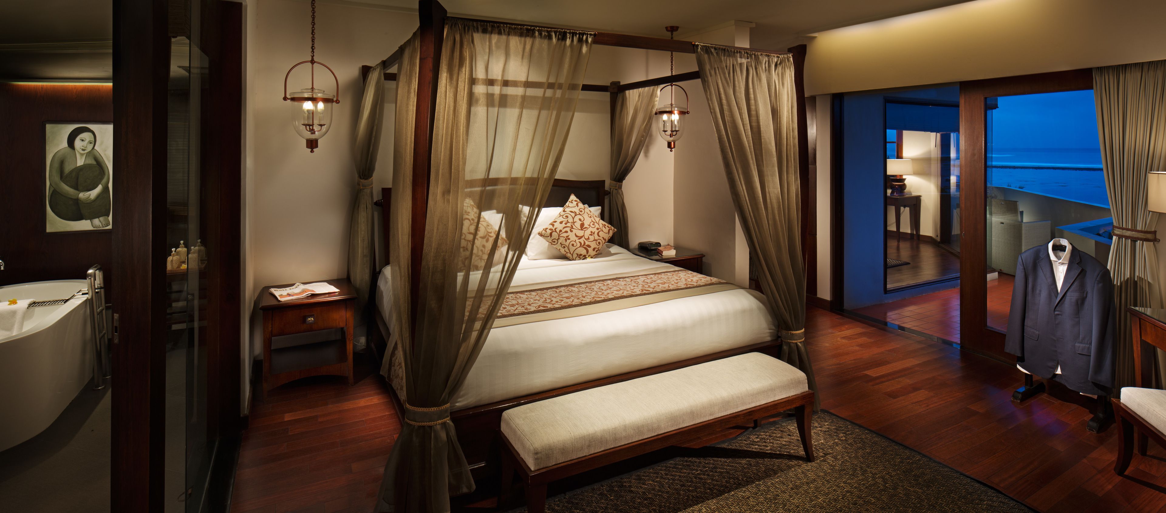 A four-poster bed with a bench, a bedside table, a nightstand, a large window with a view, and a bathroom with a bathtub and sink.