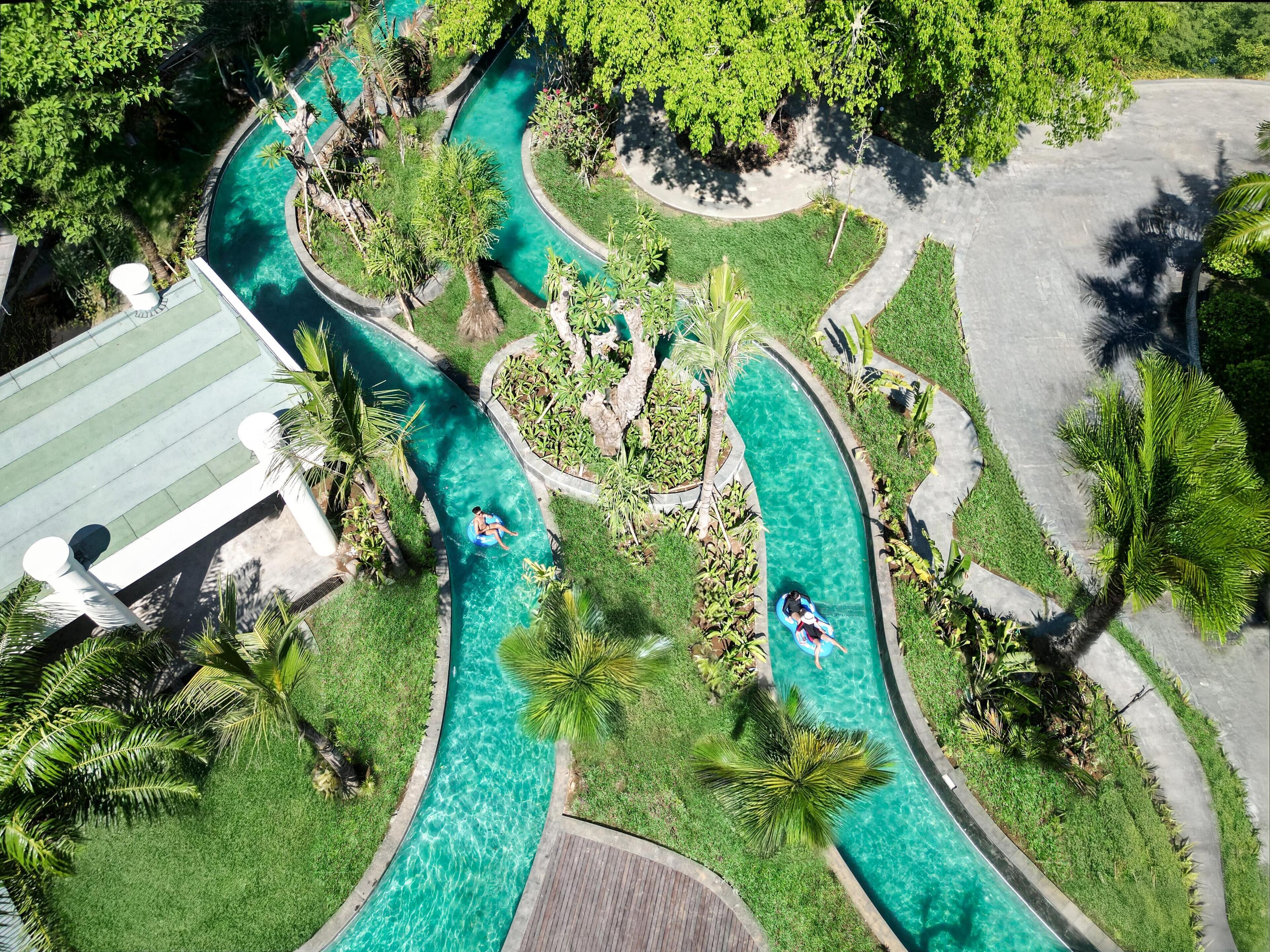 Een luchtbeeld van een resort met een slingerend zwembad, omgeven door weelderige begroeiing en palmbomen.