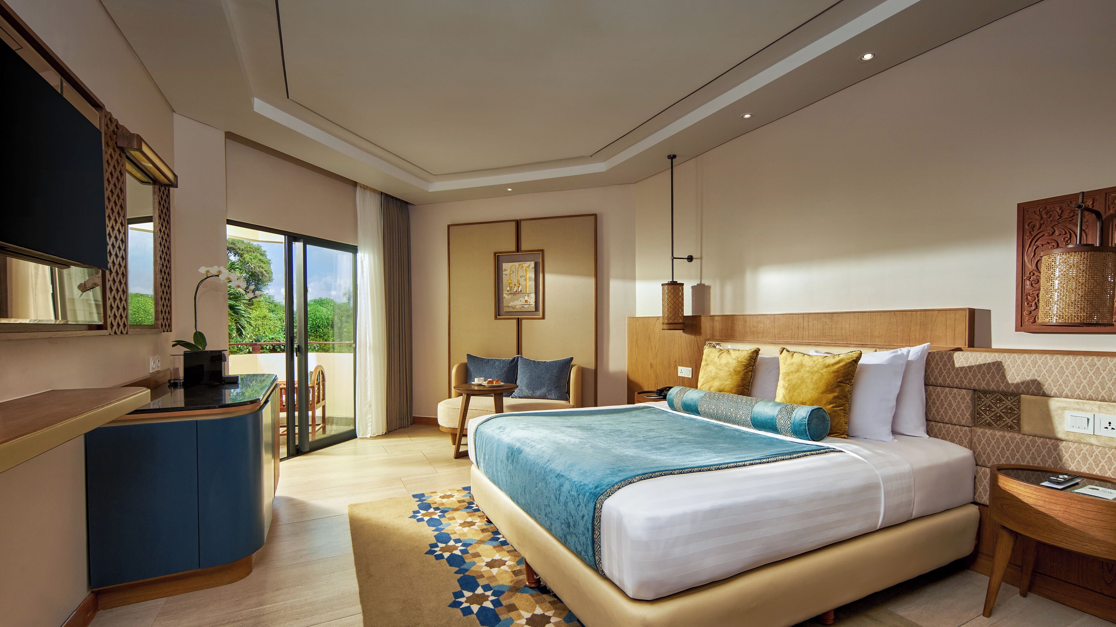 A modern hotel room with a large bed, a desk, a chair, and a balcony with a view of greenery.