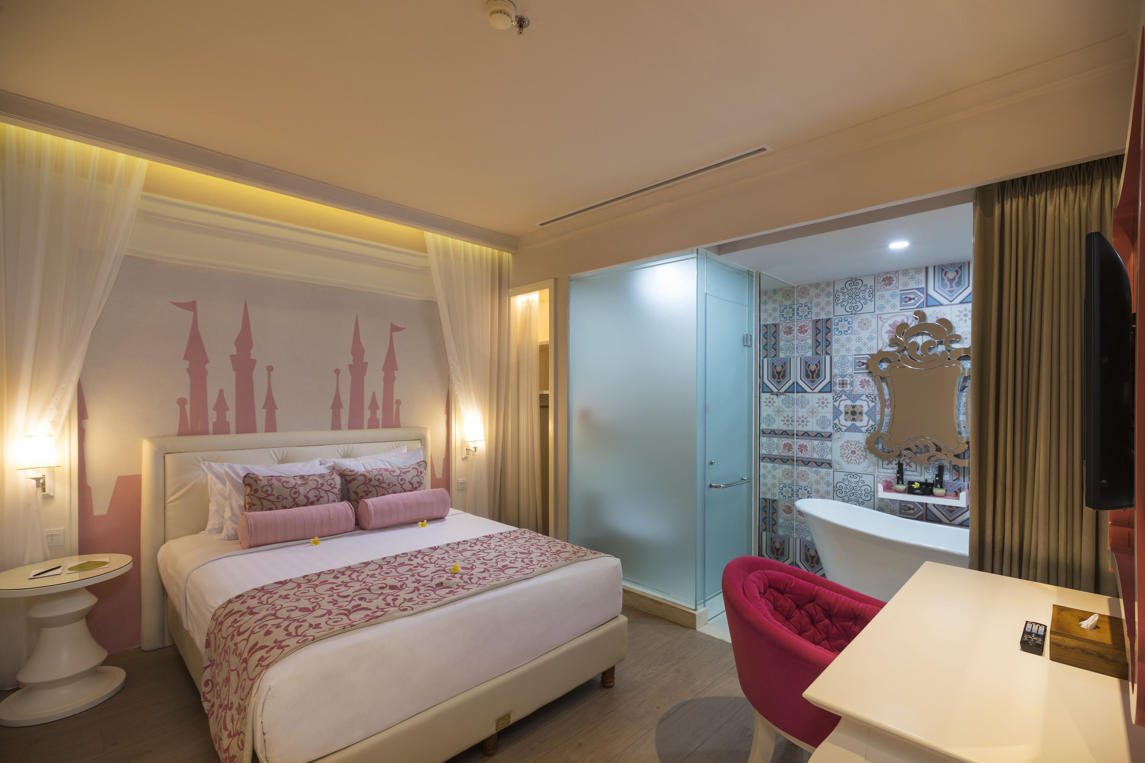 A hotel room with a bed, a vanity with a mirror, a red chair, and a small table with a remote control.
