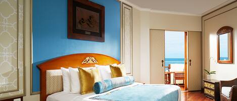 A hotel room with a large bed, a wooden headboard, a framed picture on the wall, and a view of the beach through an open door.