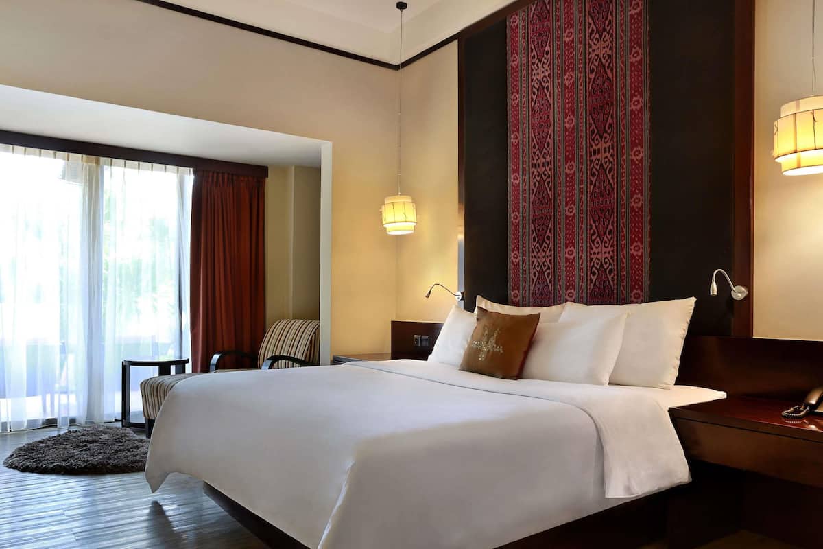 A hotel room with a large bed, a desk with a lamp, a chair, and a view of the outdoors through a window.