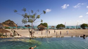 Ein Schwimmbecken mit klarem Wasser, ein Strand mit Sonnenschirmen und eine strohgedeckte Hütte.
