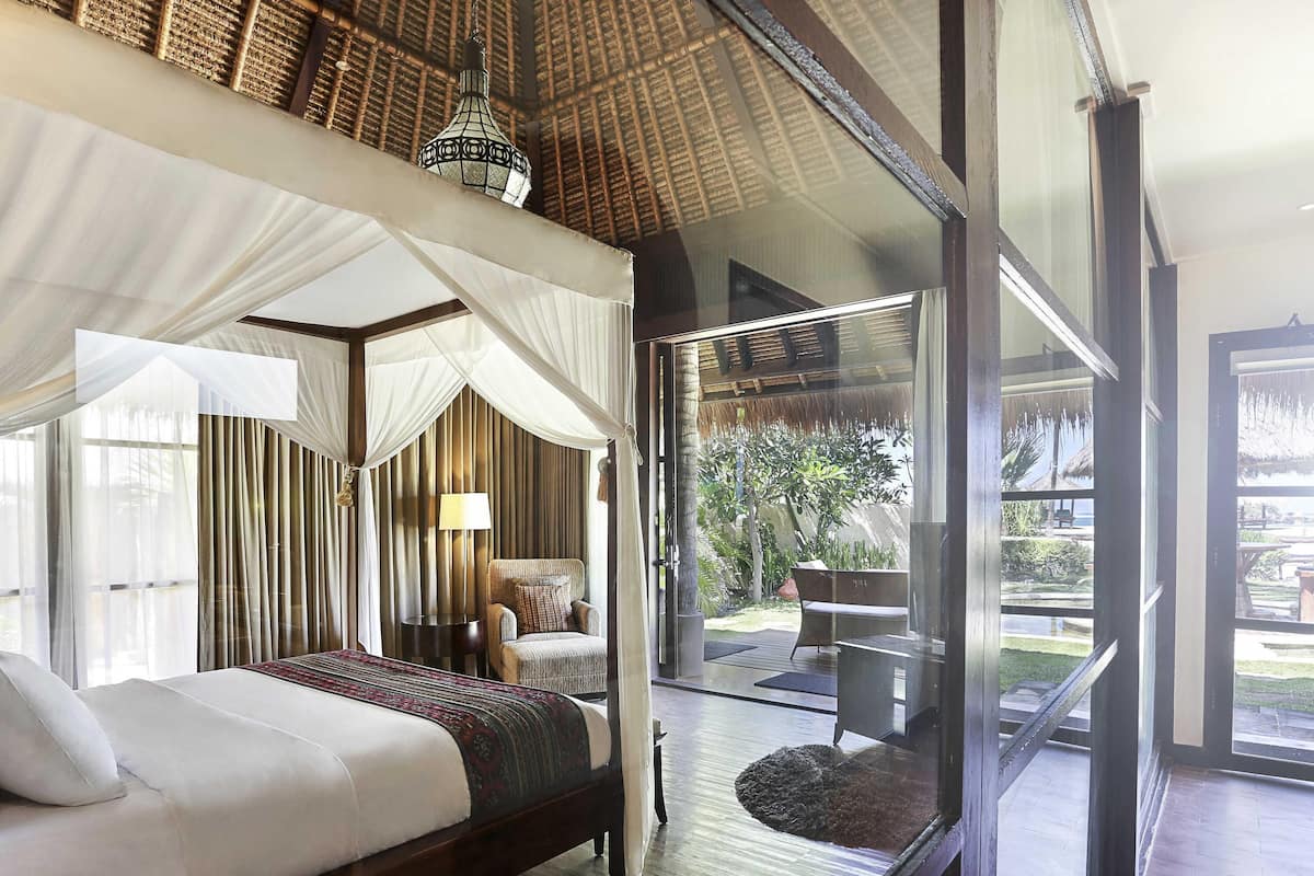 A bedroom with a bed, a nightstand, a lamp, and a view of the garden through large windows.