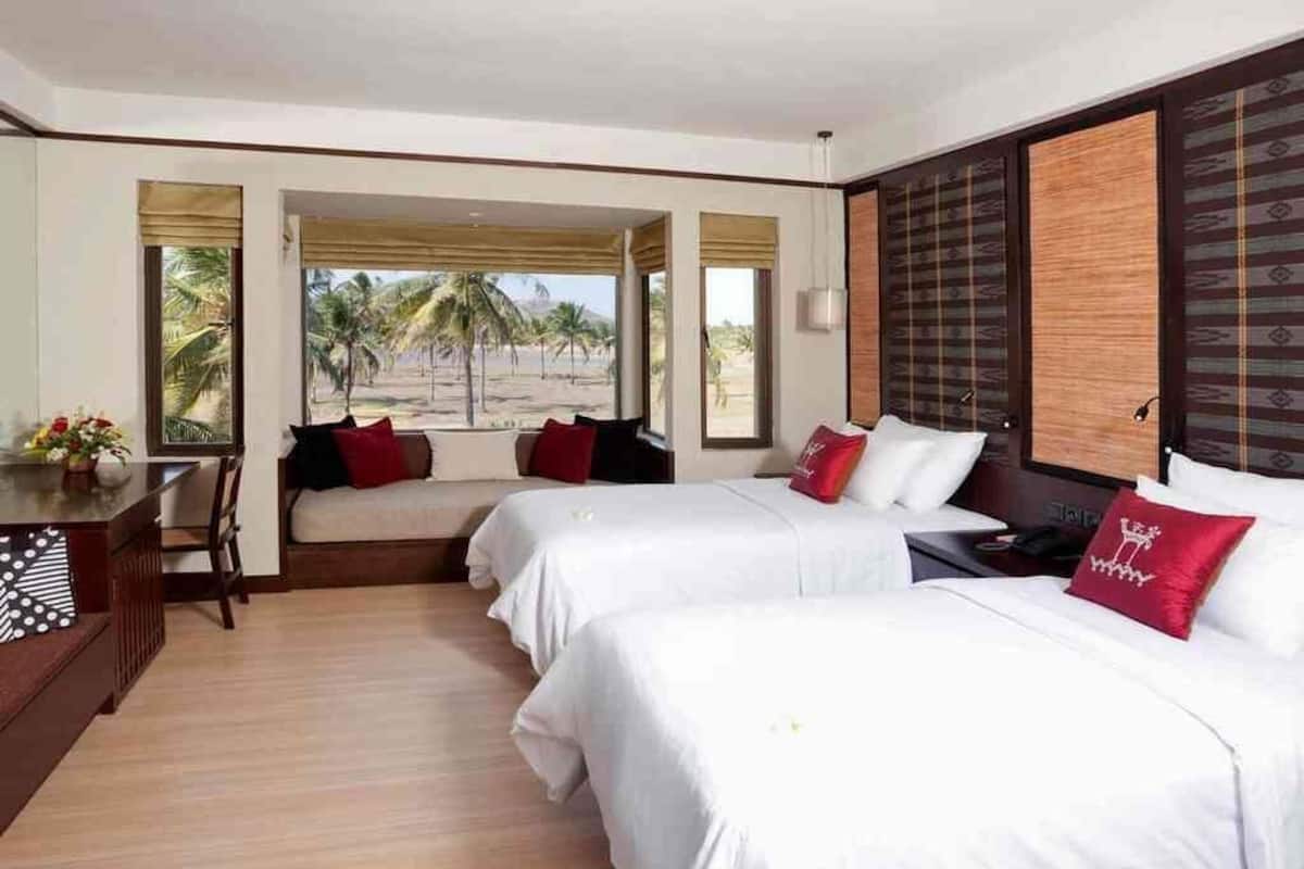 A hotel room with two beds, a desk, and a view of palm trees and mountains.