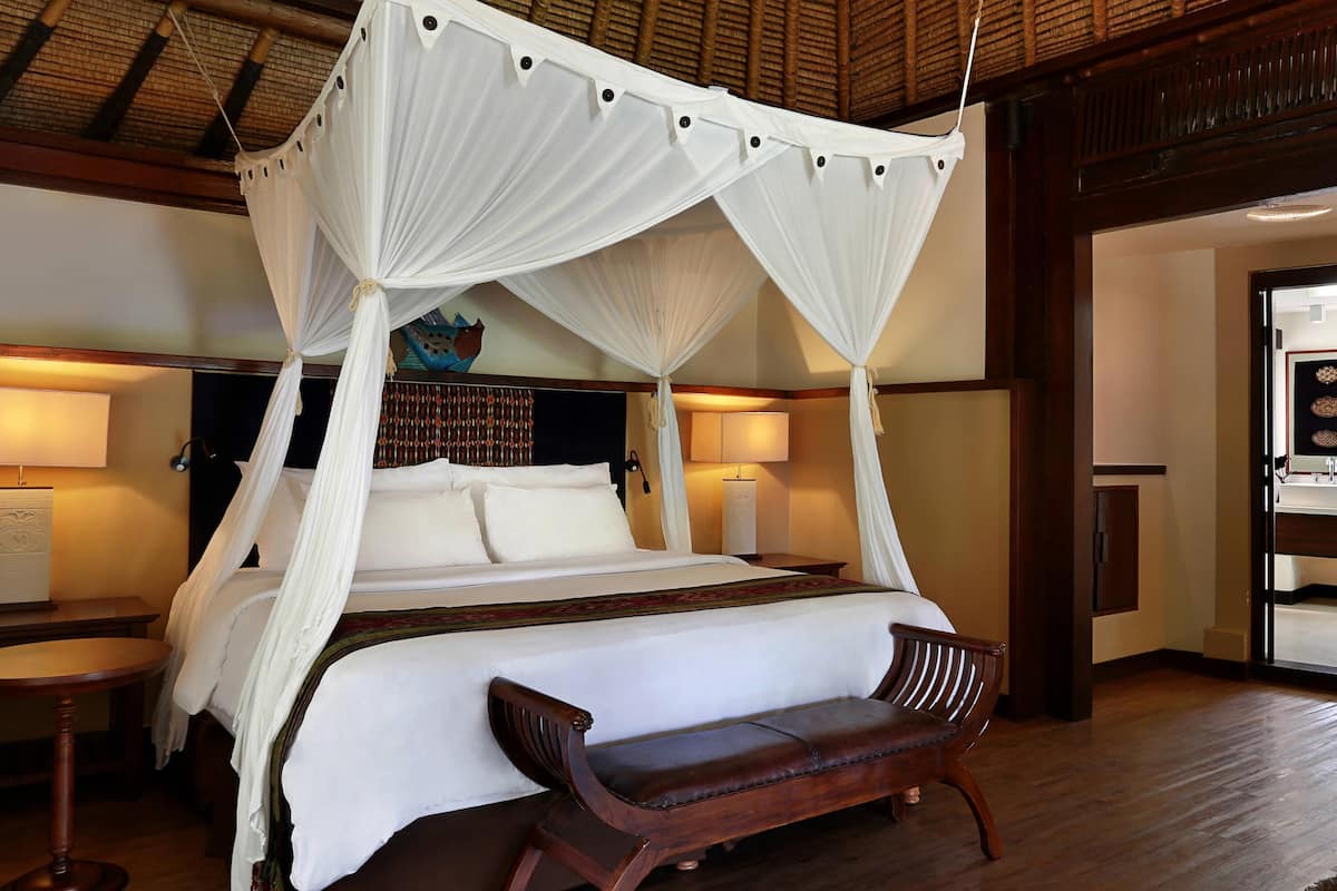 A room with a four-poster bed, a wooden bench, a round table, and a wall-mounted lamp.