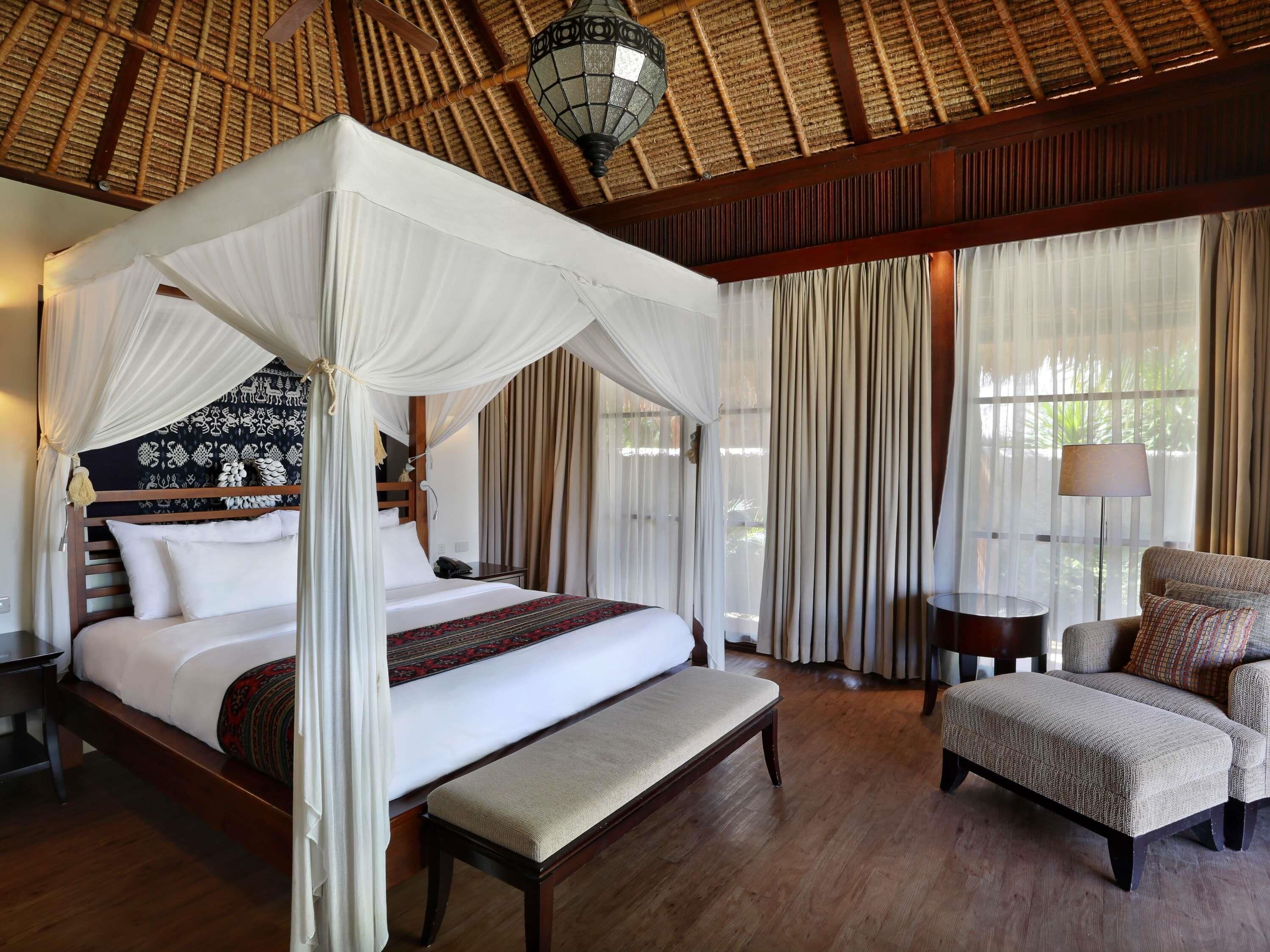 A room with a four-poster bed, a bench, a chair, a lamp, and a large window with curtains.