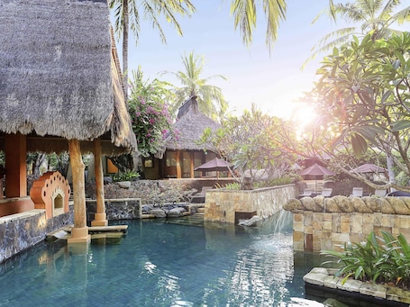 A pool area with thatched-roof structures, stone walls, and tropical plants.. Novotel Lombok Resort and Villas