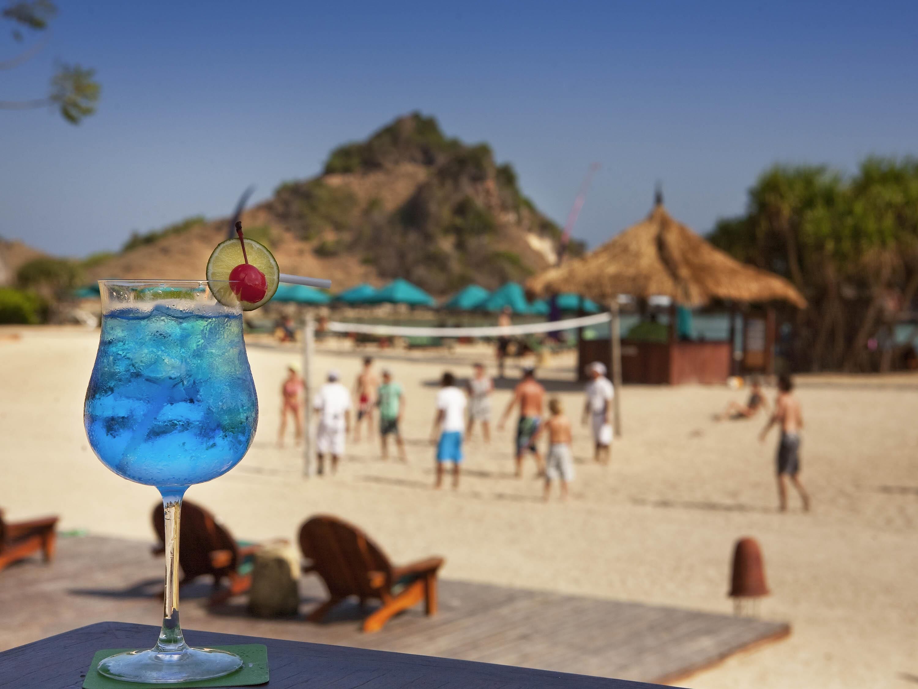 A glass of blue drink with a cherry on a stick, placed on a table with a view of a beach and people in the background.