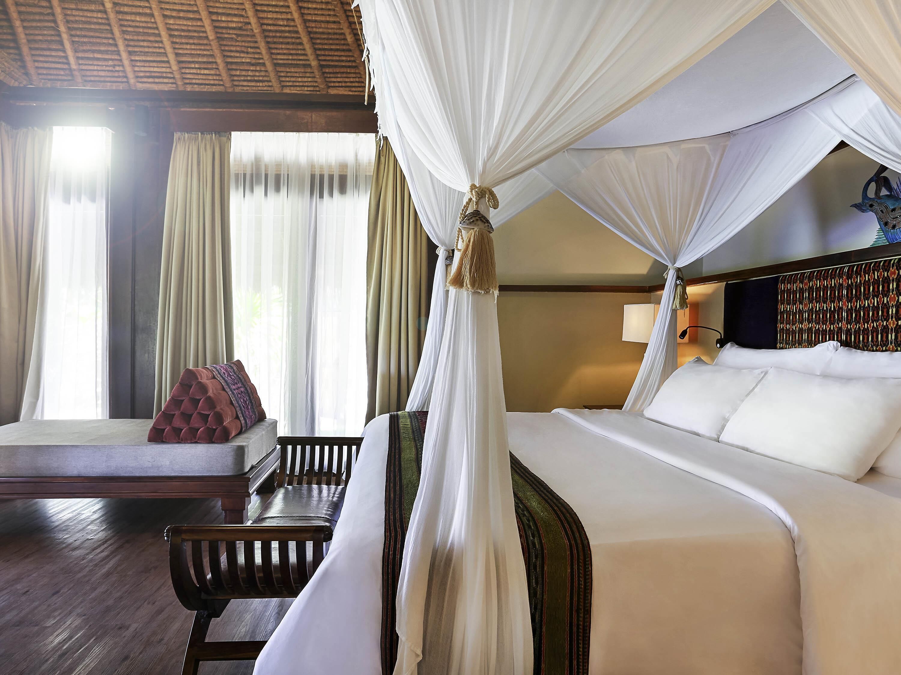 A hotel room with a bed, a bench, a window with curtains, and a canopy bed.