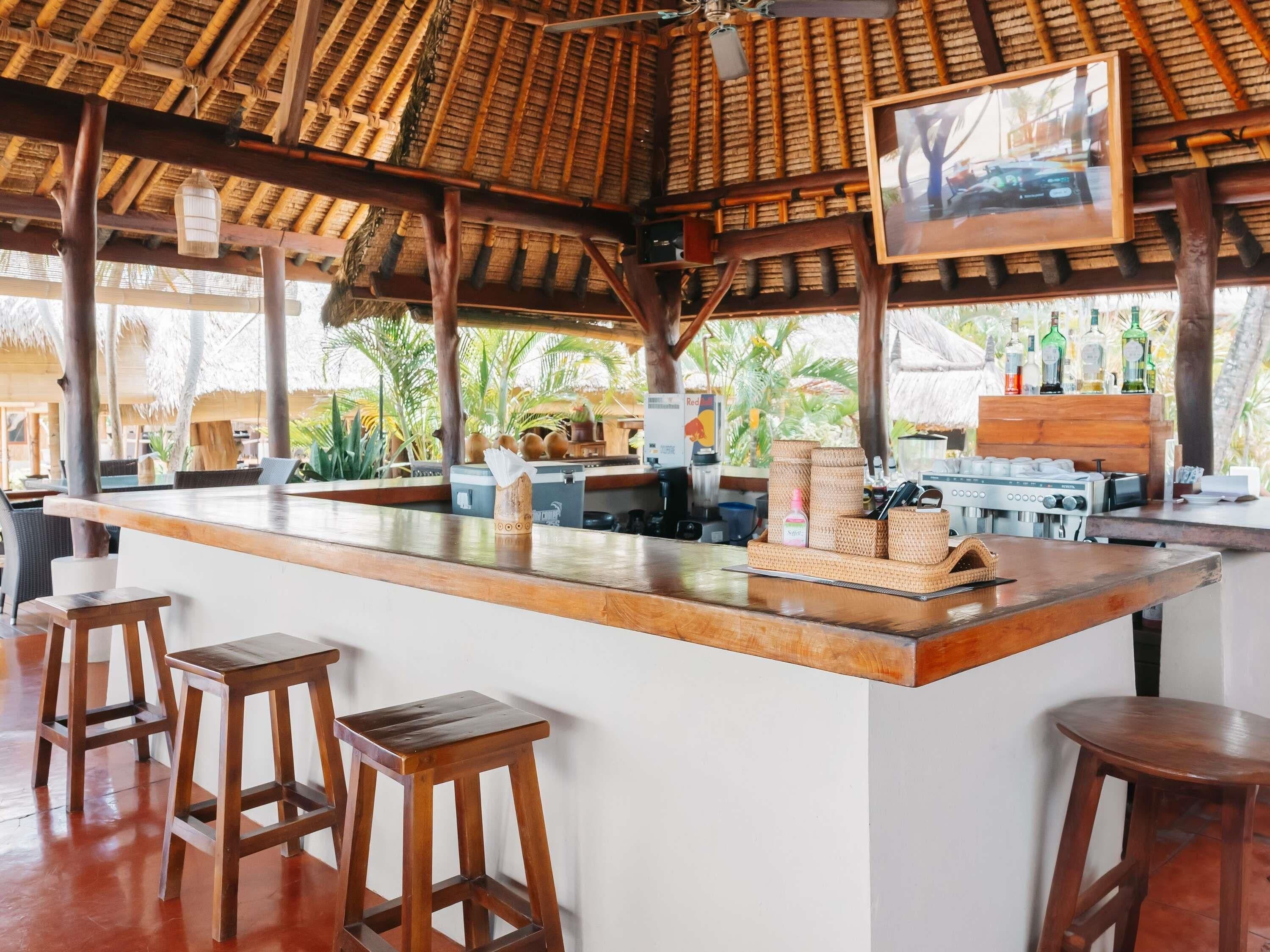 A bar area with a wooden counter, stools, and a ceiling with thatched roofing.