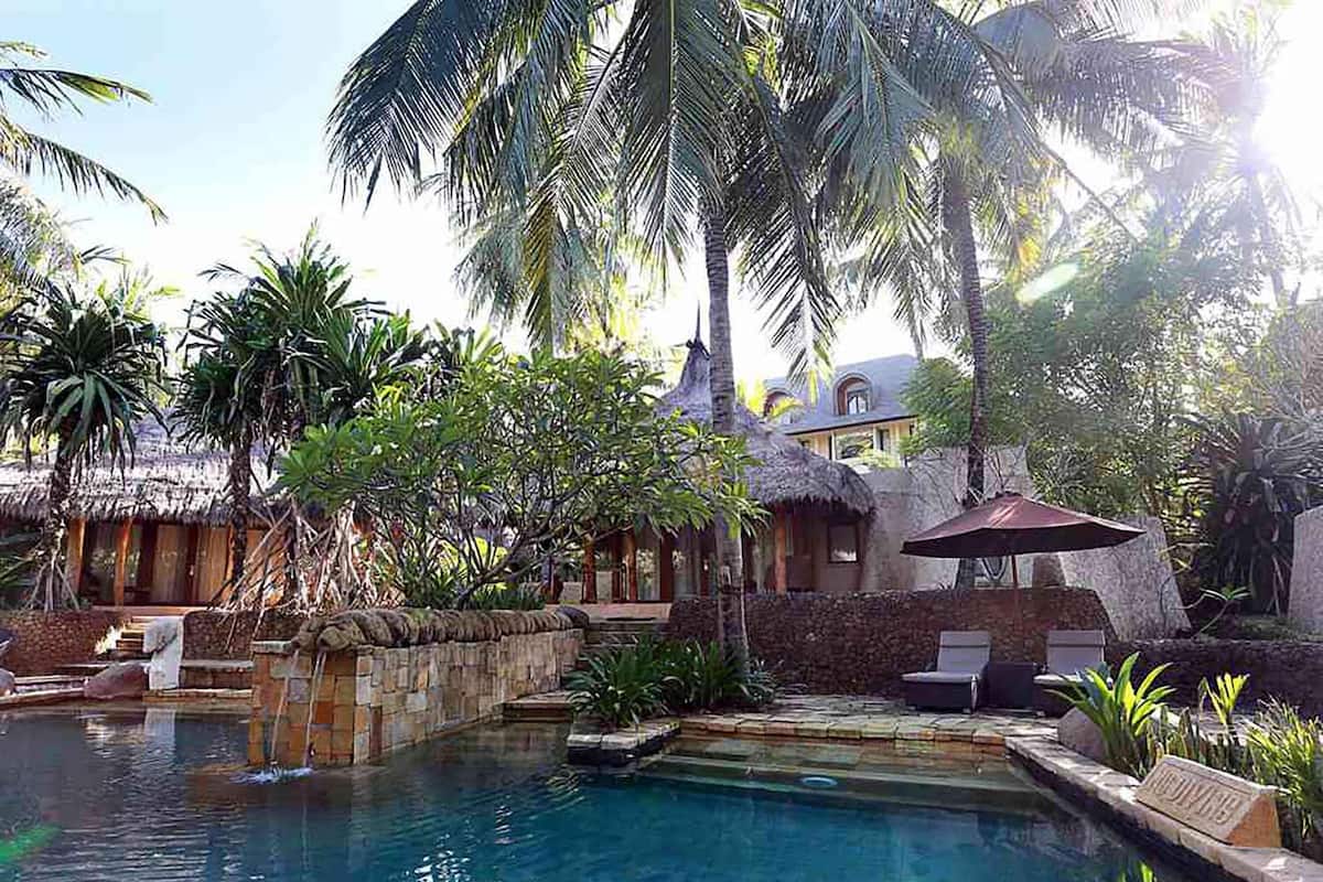 A pool area with a thatched-roof structure, lounge chairs, and tropical vegetation.