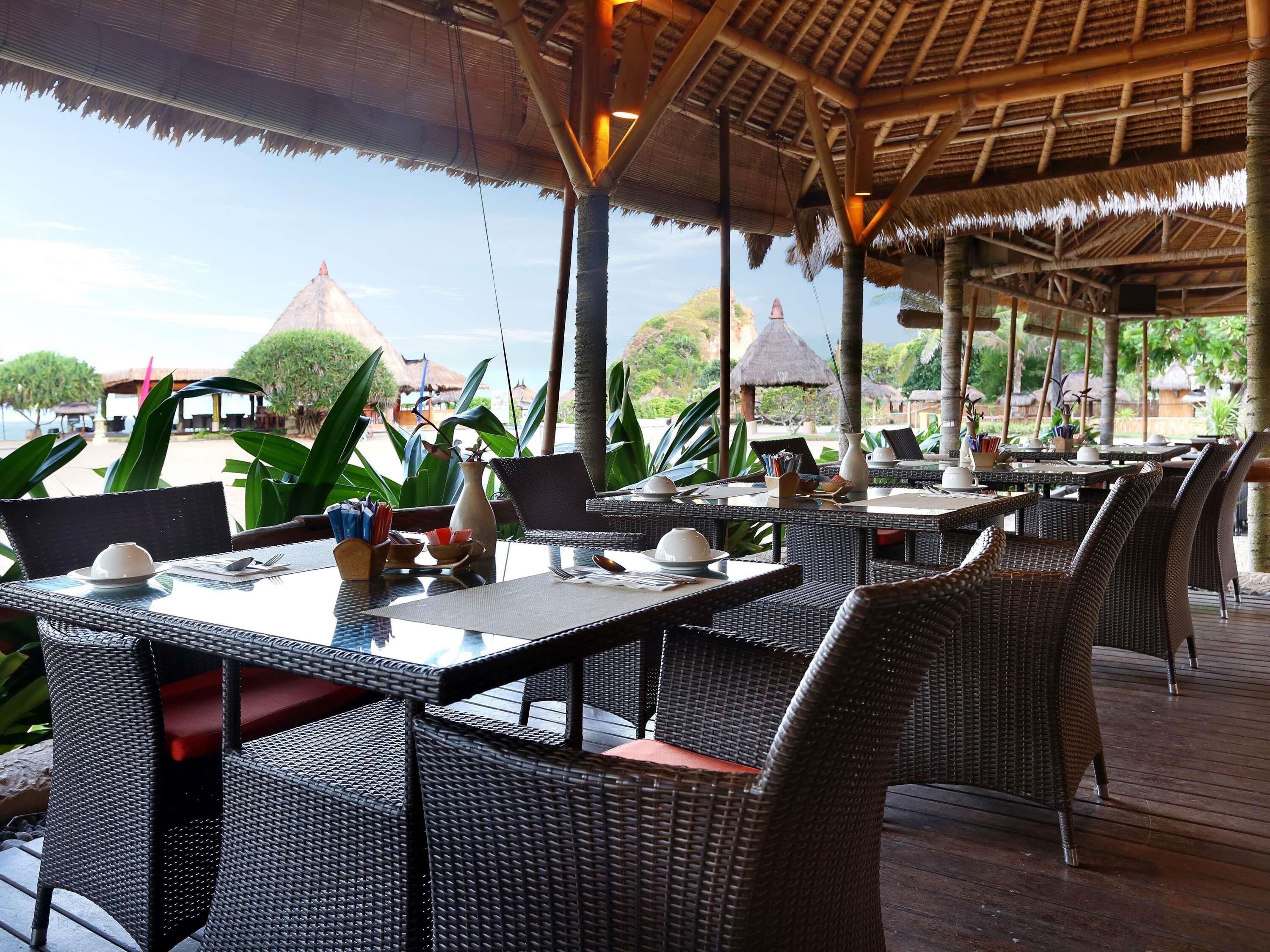 An outdoor dining area with wicker furniture, tables set with plates, cups, and cutlery, and a view of the beach and thatched-roof structures in the background.