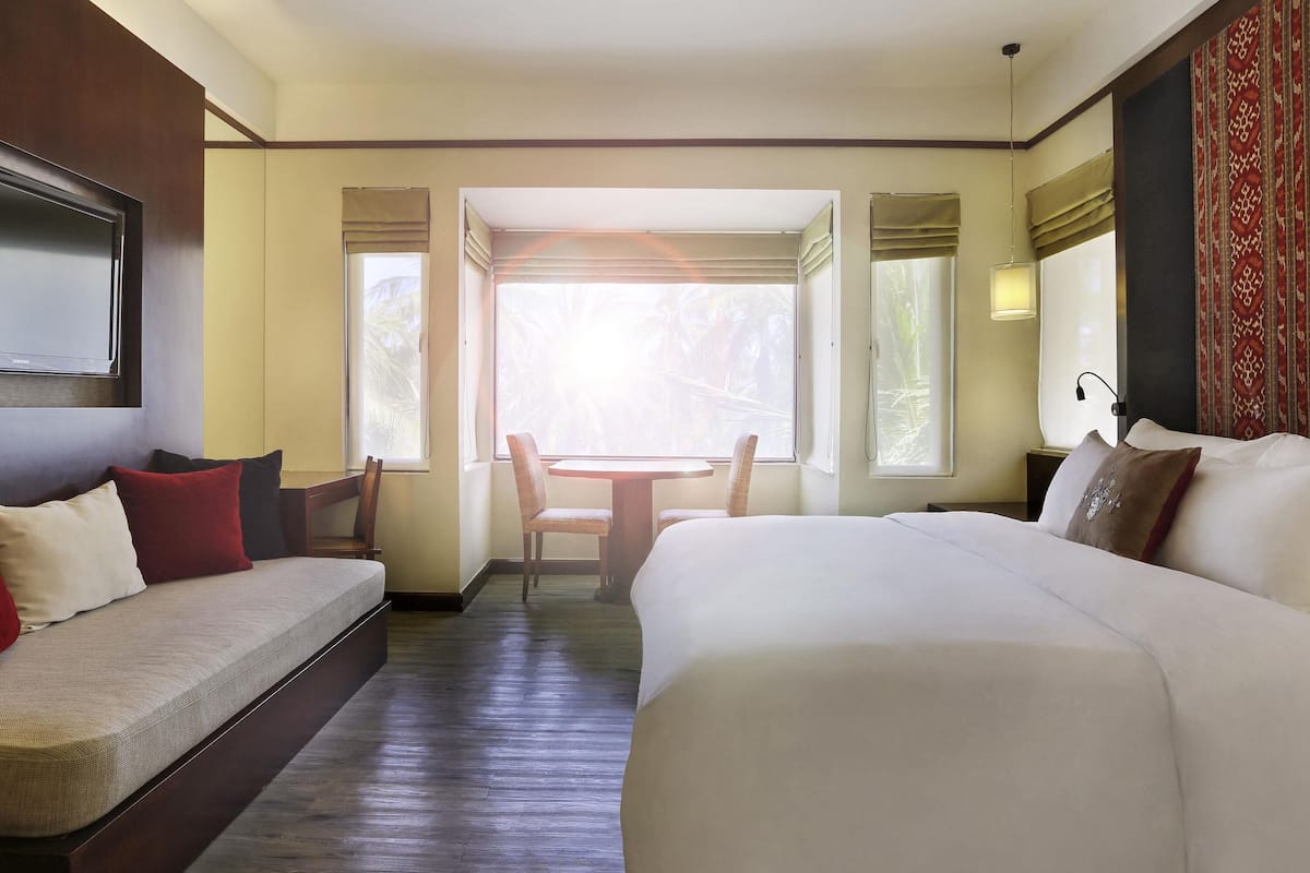 A hotel room with a large bed, a small table, and a chair. There is a window with curtains, a TV mounted on the wall, and a lamp on the nightstand.