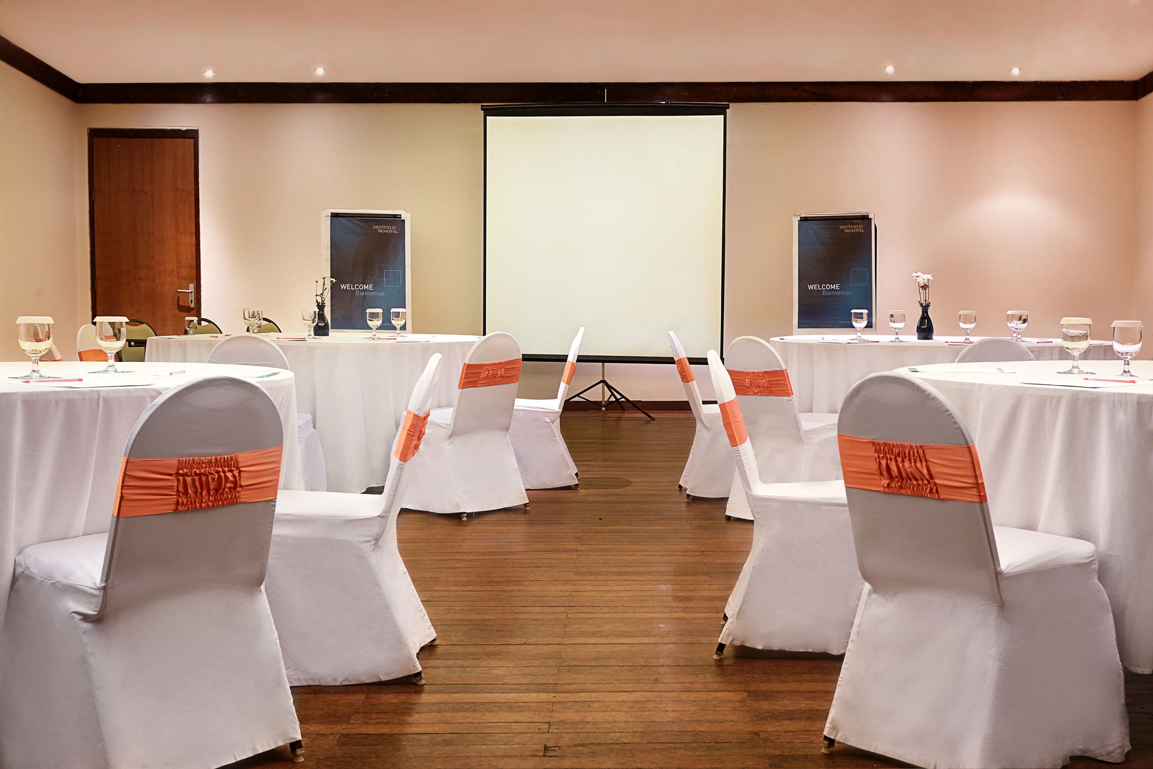 A conference room with white tables and chairs, a projector screen, and a wooden floor.