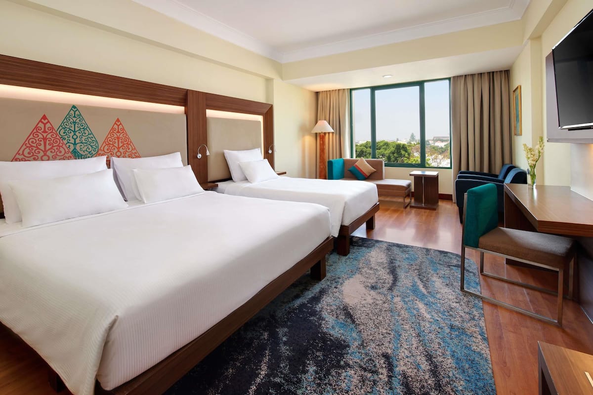A hotel room with two beds, a desk, a chair, a TV, and a large window with a view.