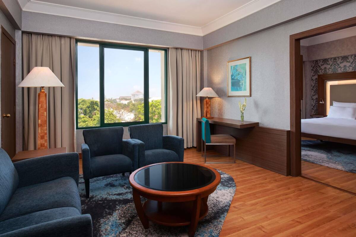A hotel room with a large window, two armchairs, a round coffee table, a bed, and a desk with a lamp and a vase of flowers.