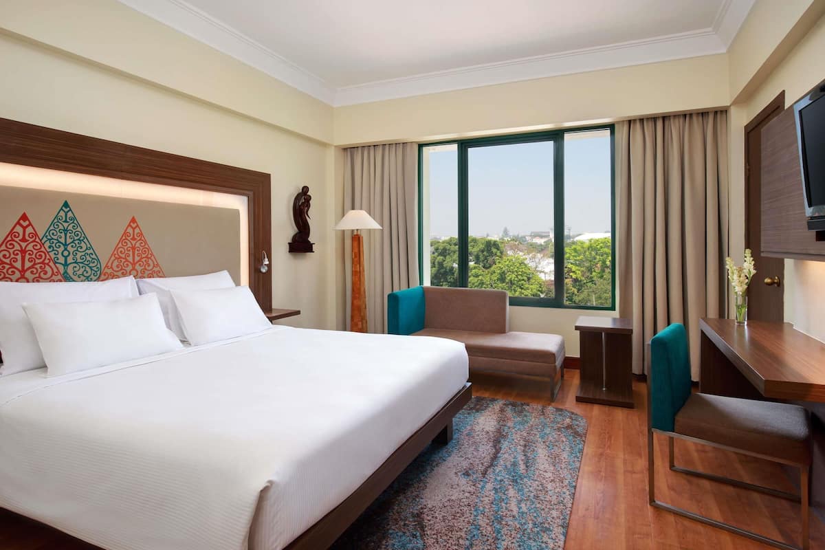 A hotel room with a large bed, a desk, a chair, a sofa, a TV, and a window with a city view.