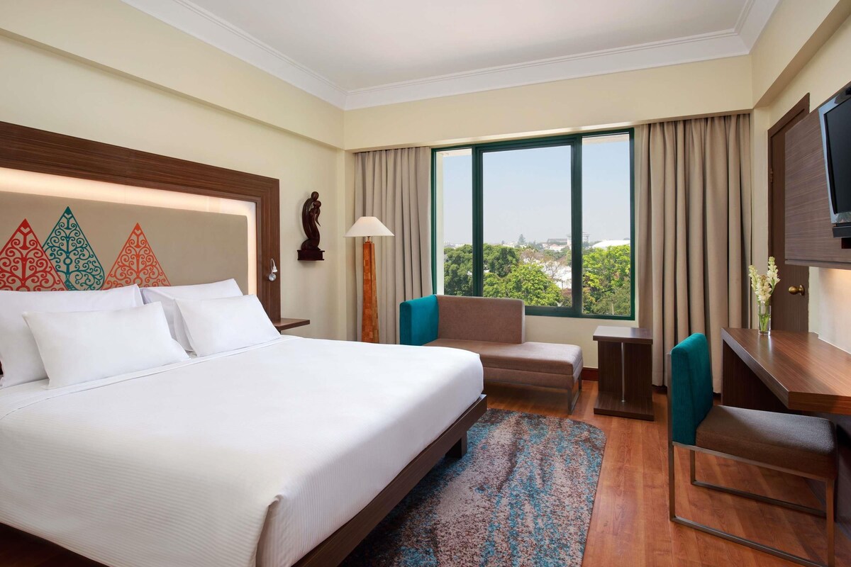A hotel room with a large bed, a desk, a chair, a sofa, a TV, and a window with a city view.