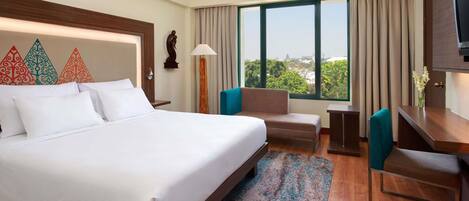 A hotel room with a large bed, a desk, a chair, a sofa, a TV, and a window with a city view.