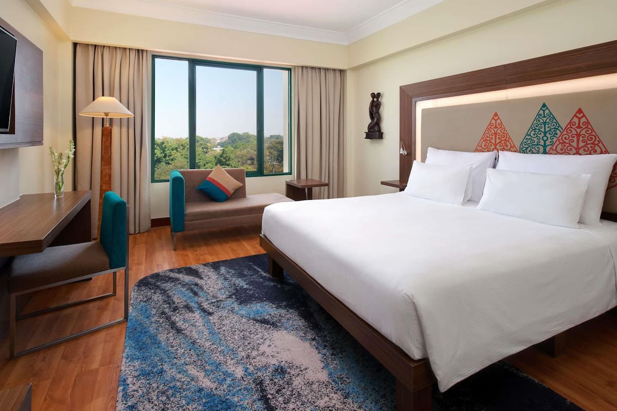 A hotel room with a large bed, a desk, a chair, a lamp, and a view of the outdoors.