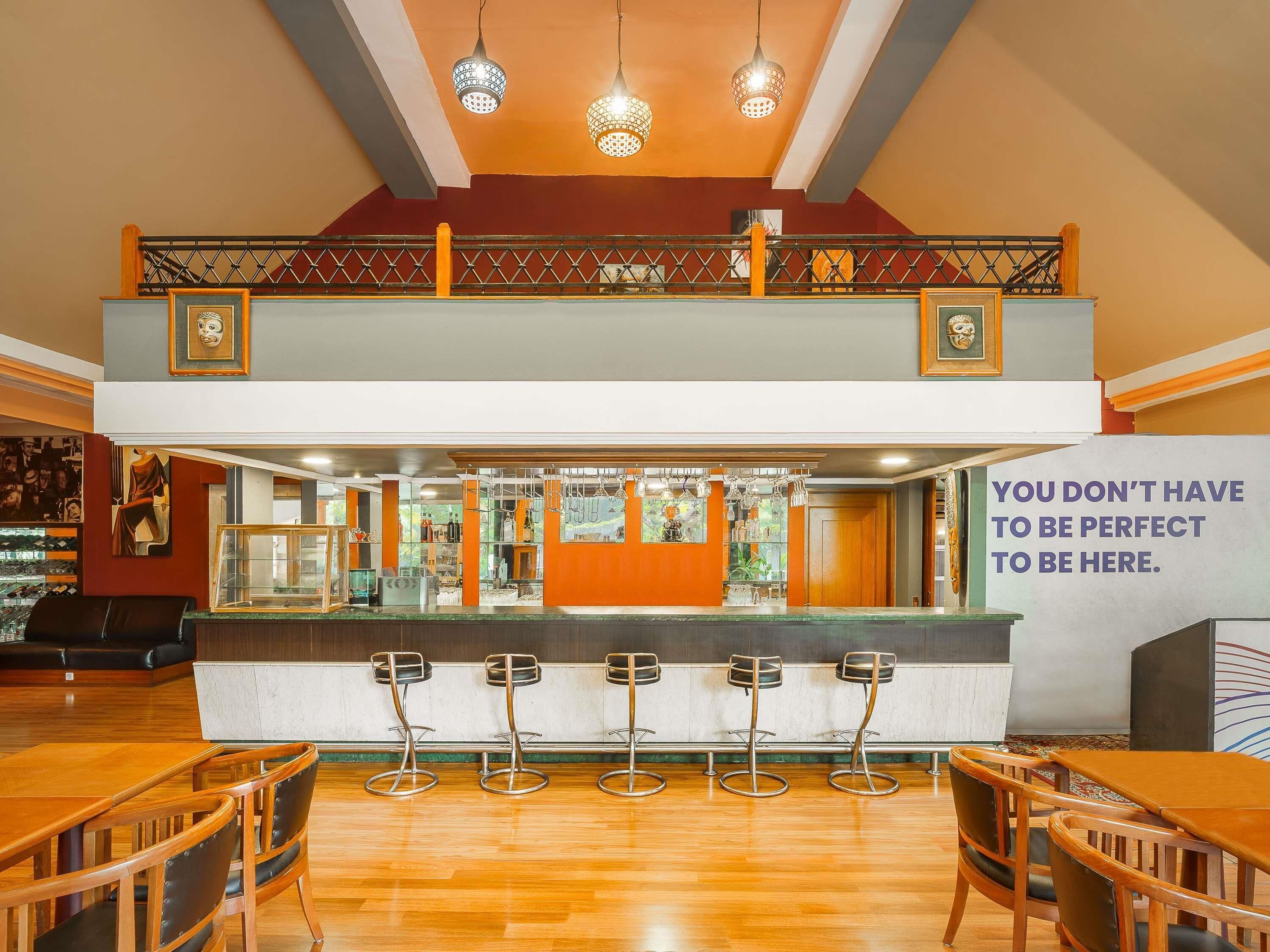A bar area with a high ceiling, multiple hanging lights, and a prominent sign stating "YOU DON'T HAVE TO BE PERFECT TO BE HERE."