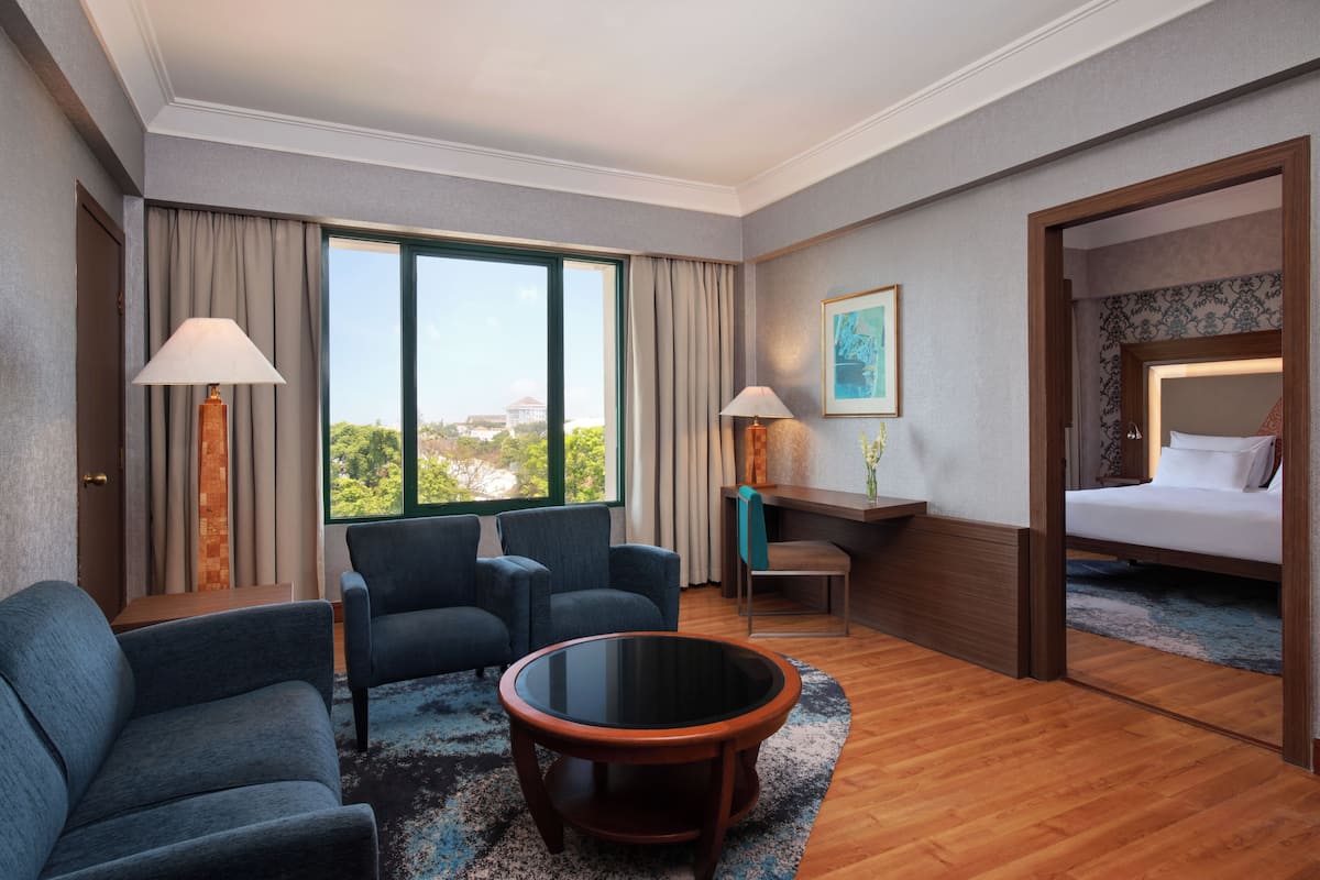 A hotel room with a bed, two armchairs, a coffee table, a desk, two lamps, and a large window with a view.