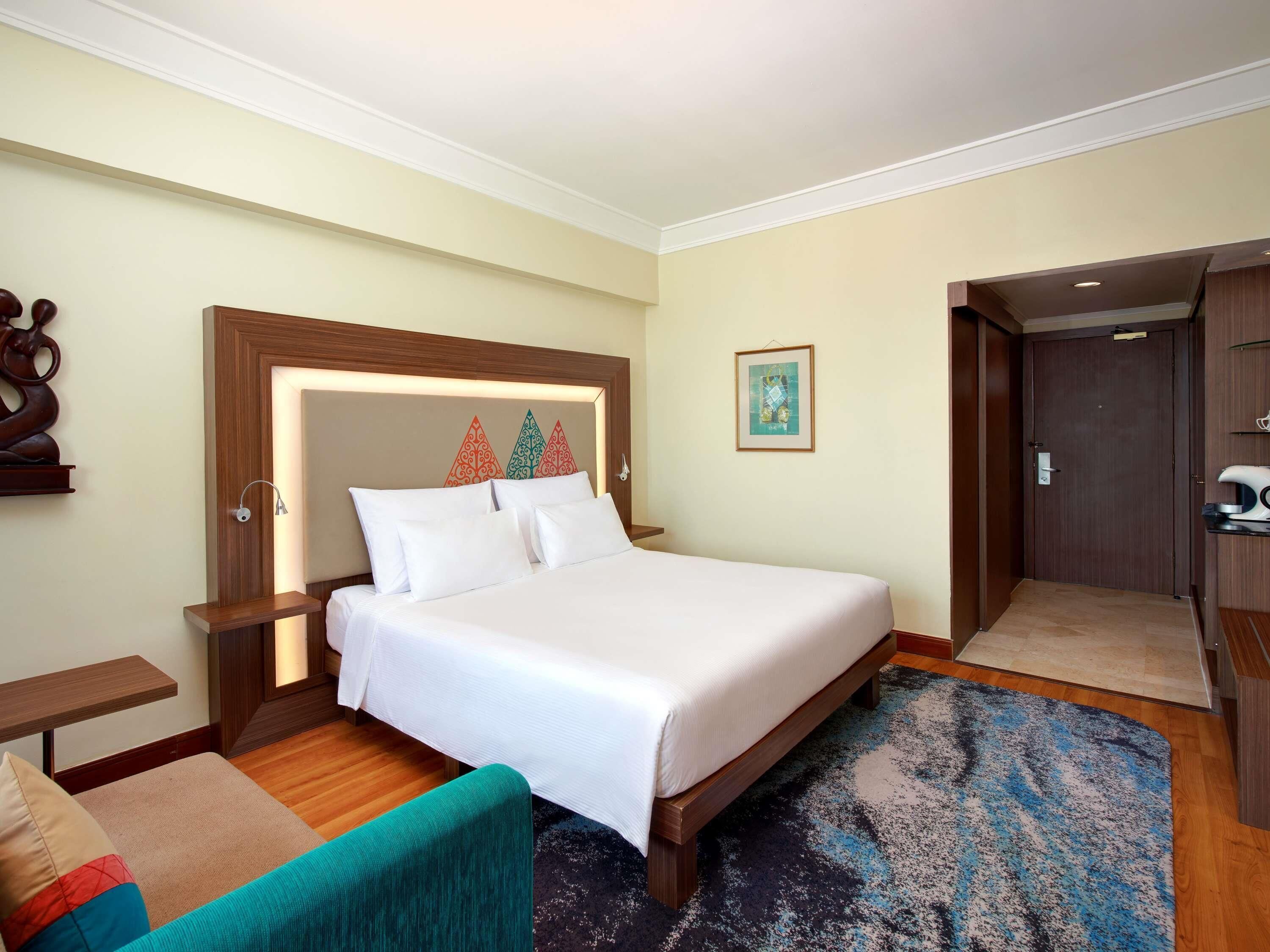 A hotel room with a large bed, a wooden headboard, a bedside table, a chair, a colorful rug, and a door leading to a bathroom.