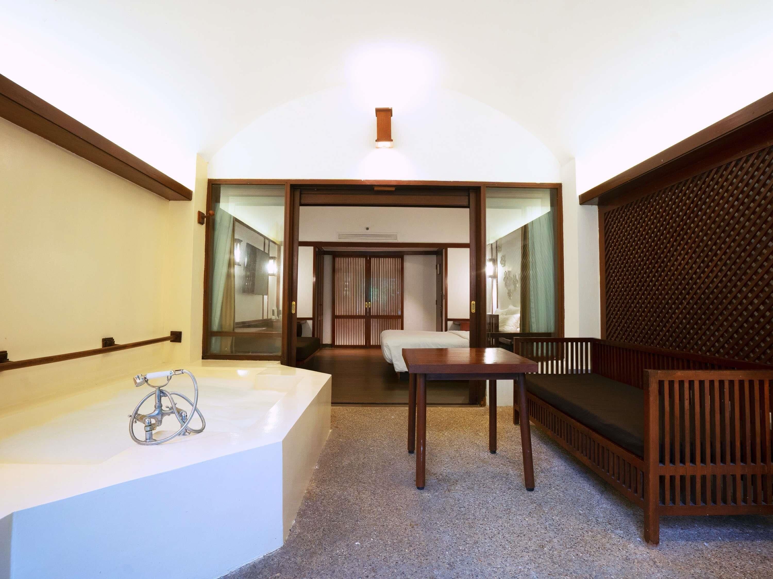 A hotel room with a bathtub, a wooden table, a bench, and a sliding door leading to a bedroom.