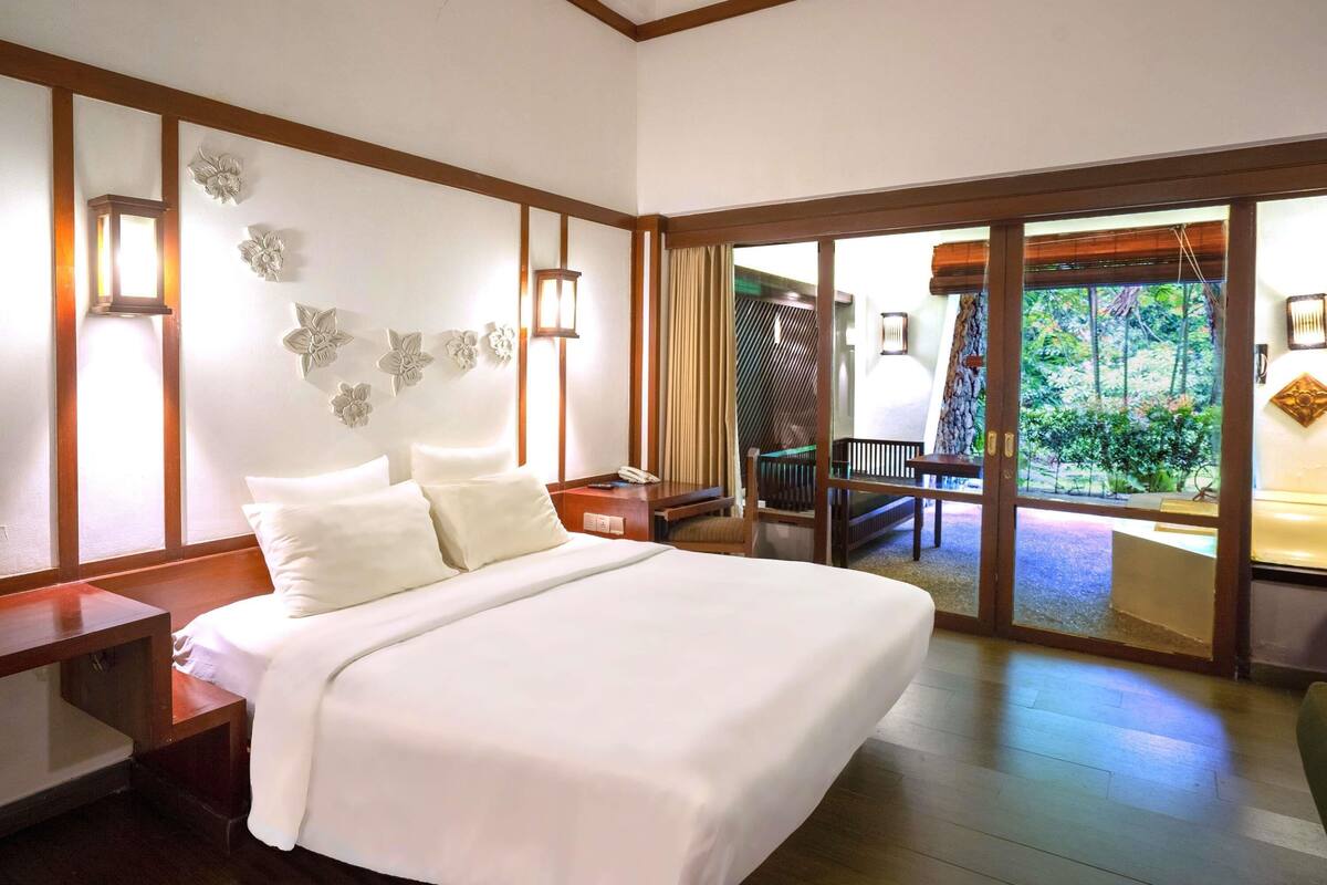A bedroom with a large bed, a nightstand, a desk, and a view of a balcony with outdoor furniture.