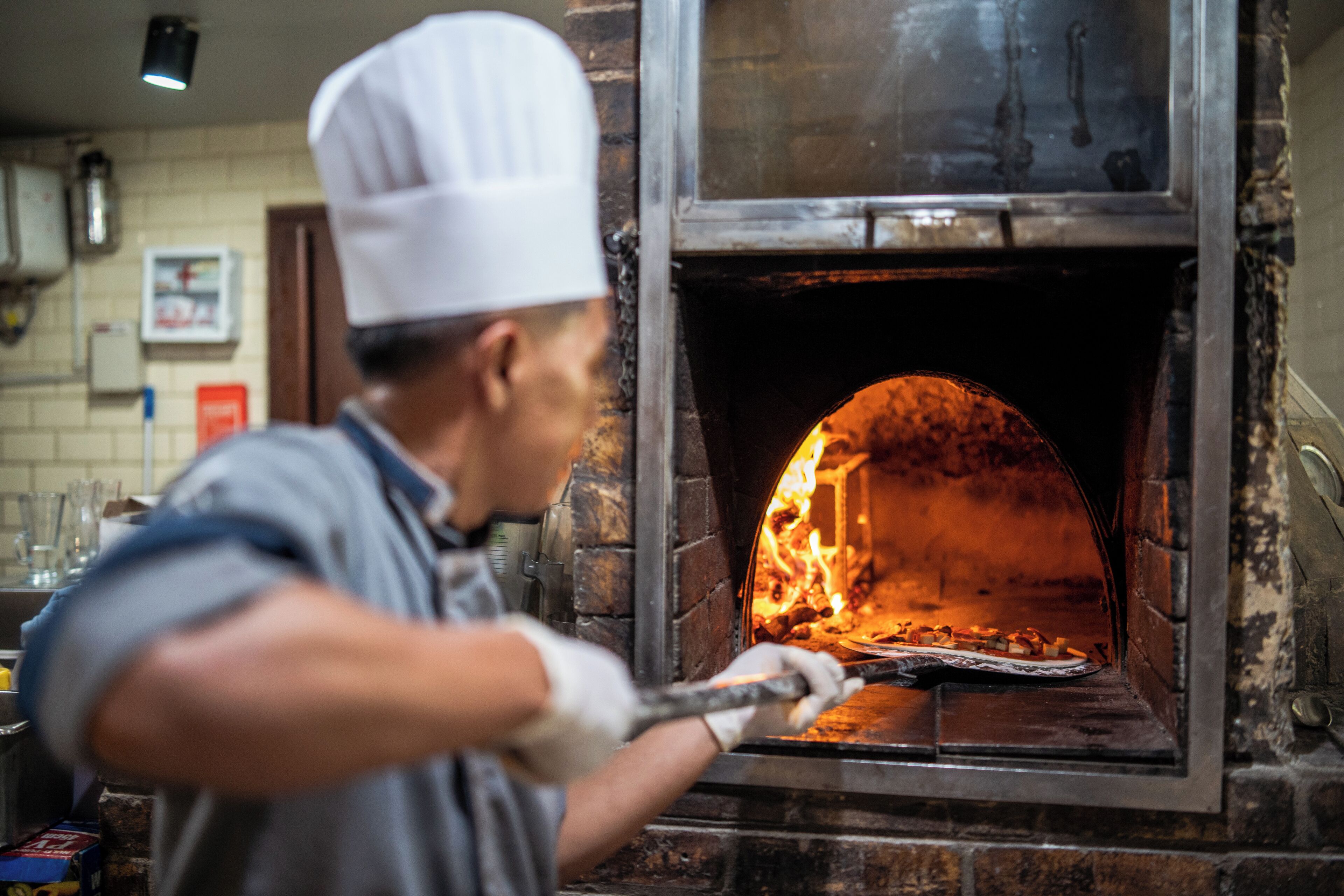 A chef in a white hat and apron using a long-handled tool to manage a pizza inside a brick oven.