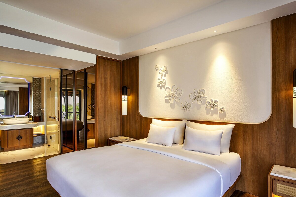 A modern hotel room with a large bed, a bathroom with a glass shower enclosure, and a wooden headboard with wall-mounted lights.