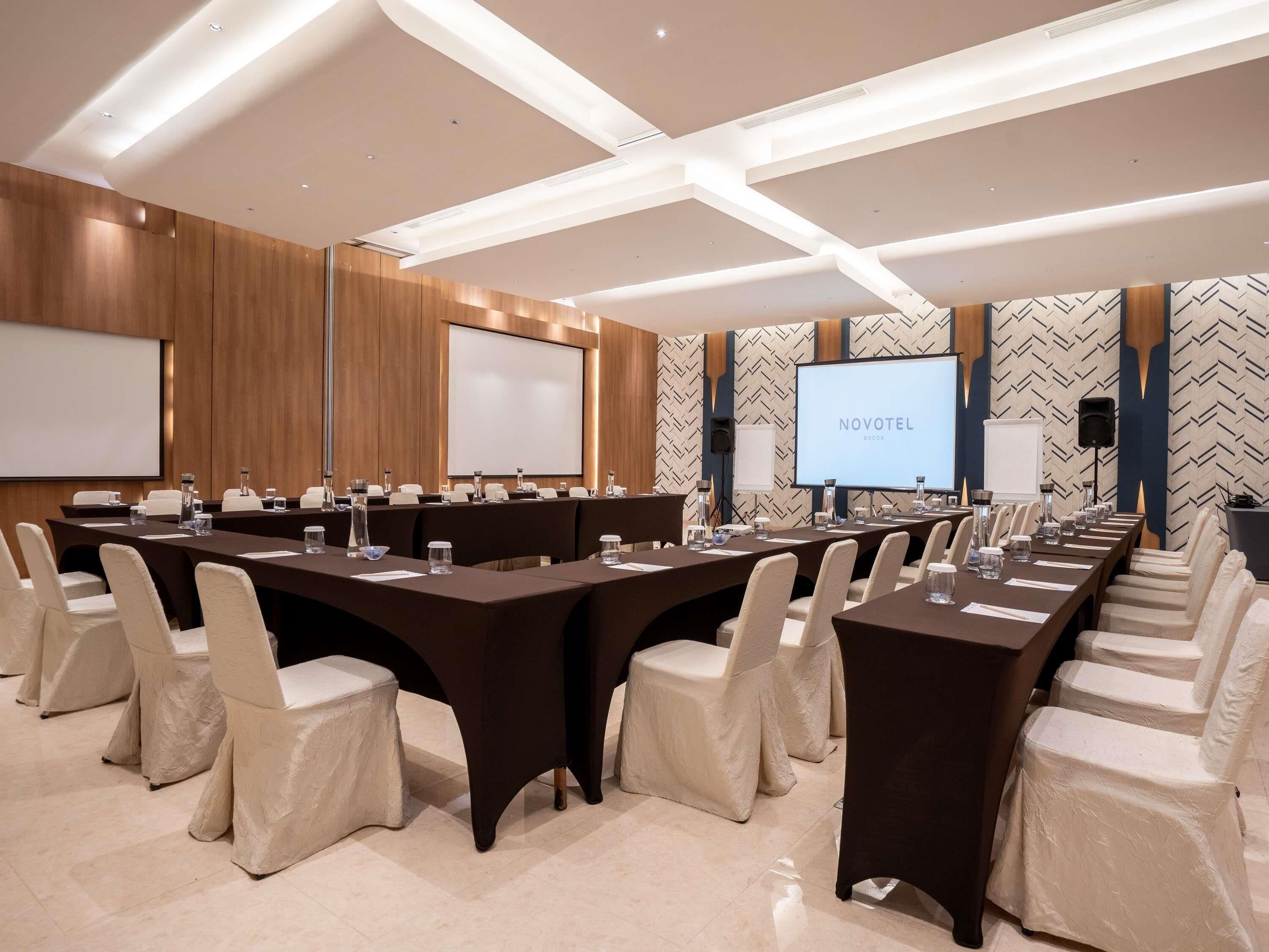 Una sala de conferències amb una taula llarga, cadires, una pantalla de projector i un cartell de 'NOVOTEL'.