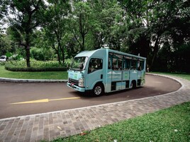 Un autobús azul claro con la marca 'NOVOTEL' estacionado en una calle con bordillo de ladrillo, rodeado de vegetación.