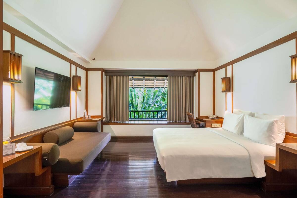 A hotel room with a bed, a sofa, a TV, a window with curtains, and a balcony view.