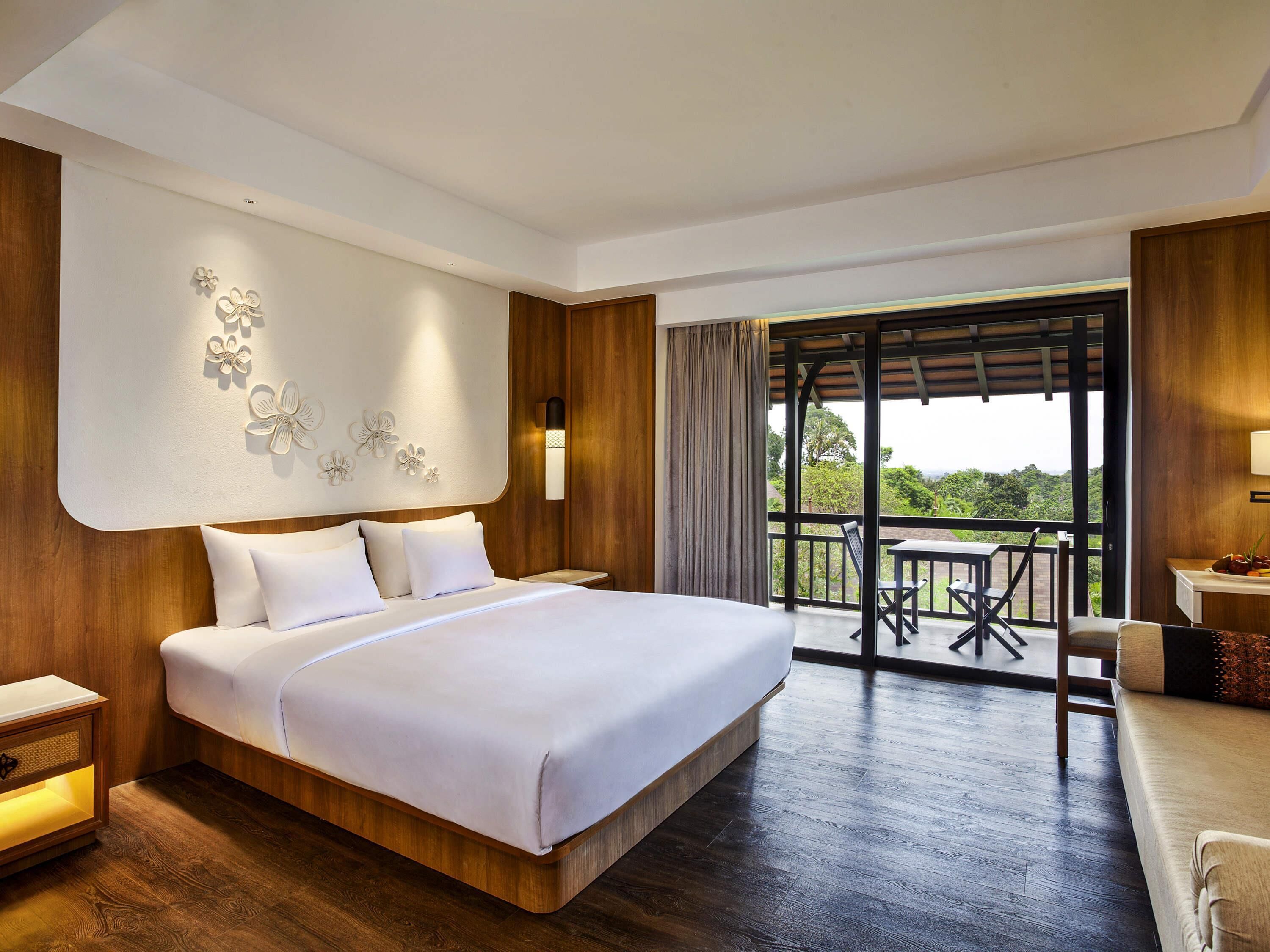 Una habitación de hotel moderna con una cama grande, paneles de madera, un balcón con mesa y sillas, y vistas al exterior.