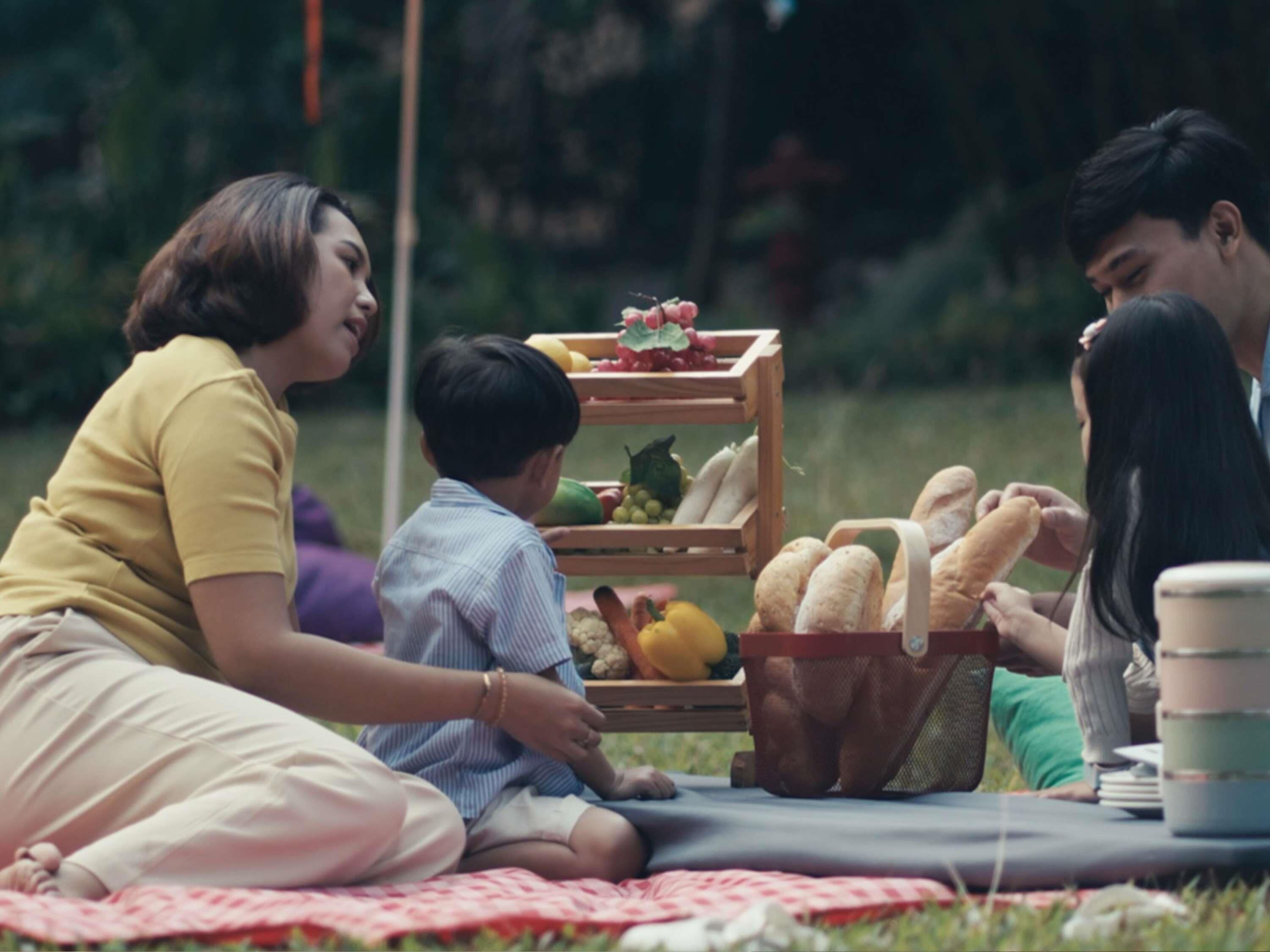 Un montaje para picnic con una manta, una cesta de frutas y verduras, y una cesta de pan sobre el césped.