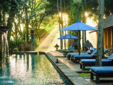 A serene pool area with lounge chairs and umbrellas, surrounded by lush greenery and a stone pathway.. Novotel Bogor Golf Resort & Convention Center