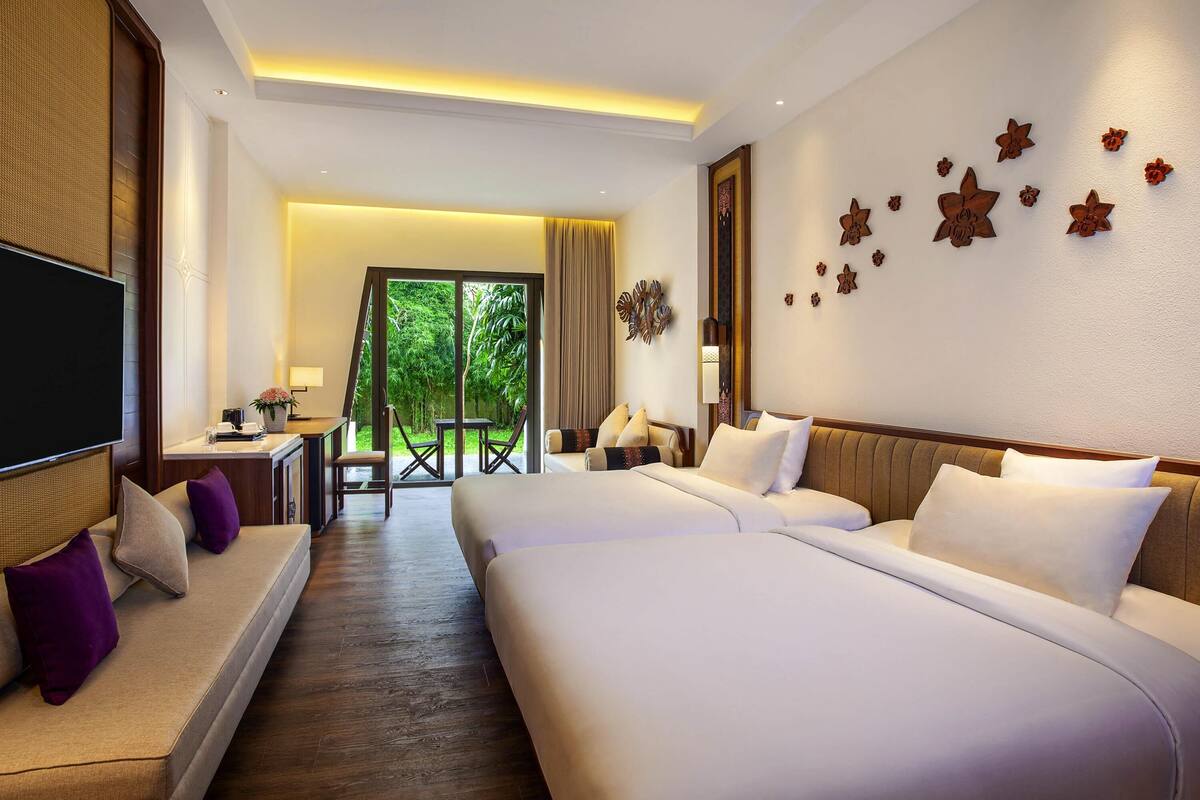 A hotel room with a large bed, a TV, a desk with a chair, and a view of greenery through the window.