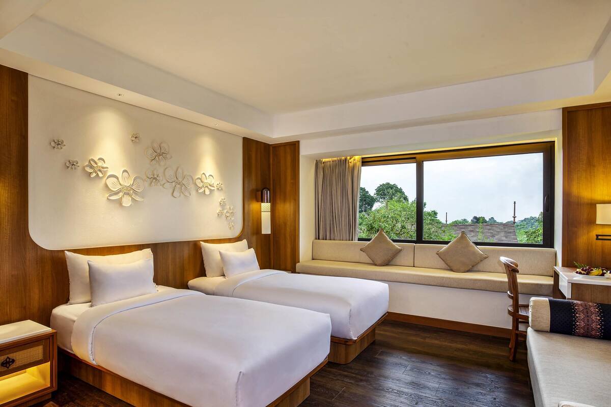A hotel room with two beds, a sofa, a window with a view, and a wall-mounted light fixture.