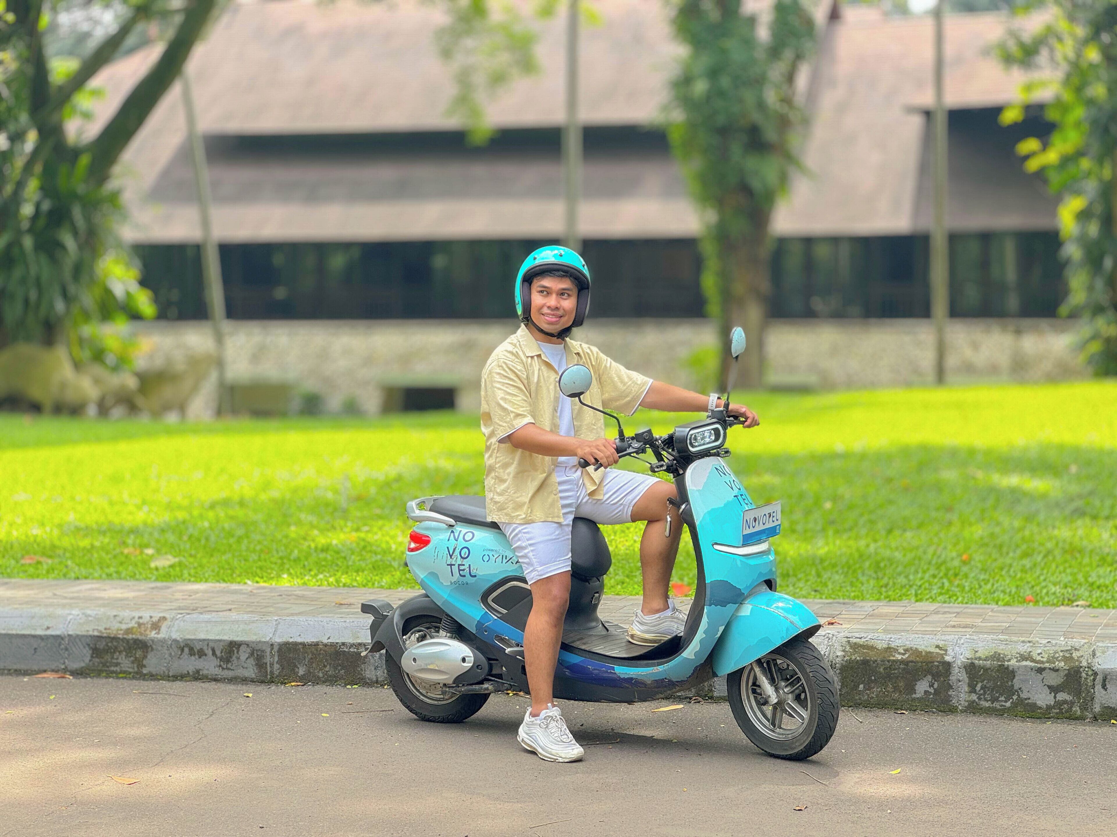A person sitting on a scooter in a park with a building in the background.