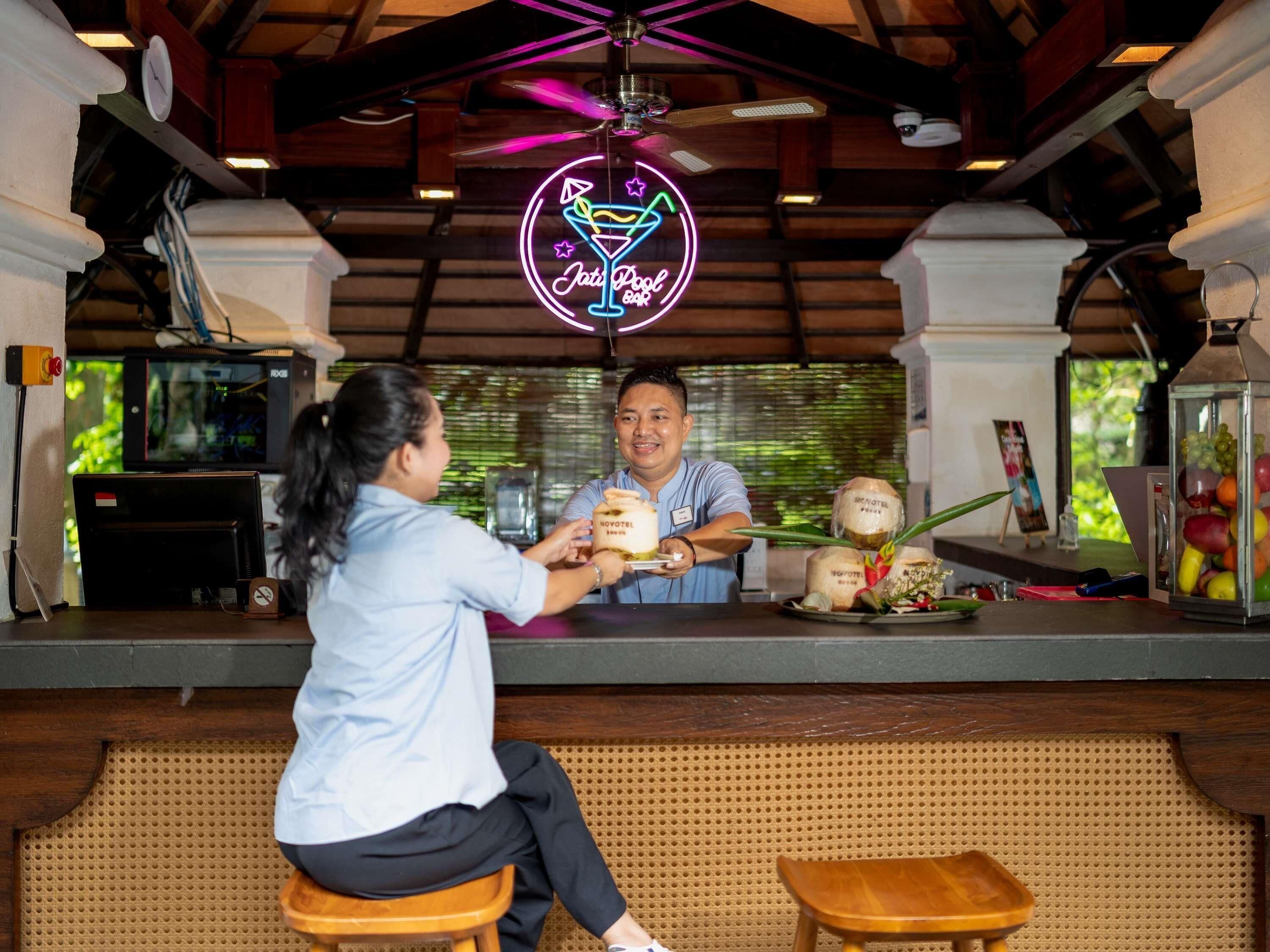 A bar area with a neon sign, a ceiling fan, and a counter with a display of fruits and a cake.
